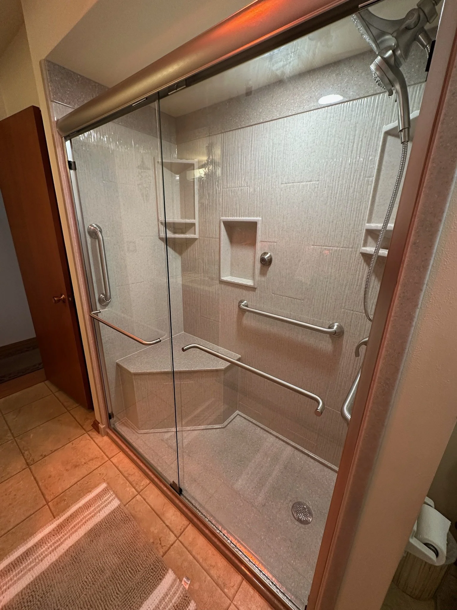 A walk-in shower with glass doors, metal grab bars, built-in shelves, and a handheld showerhead inside a tiled bathroom.