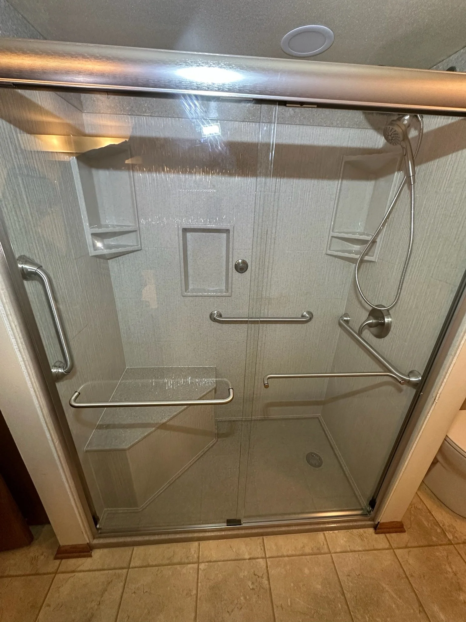 A clear shower enclosure with sliding glass doors, metal handles, a built-in corner seat, and multiple grab bars, with a showerhead on a flexible hose attached to the wall.