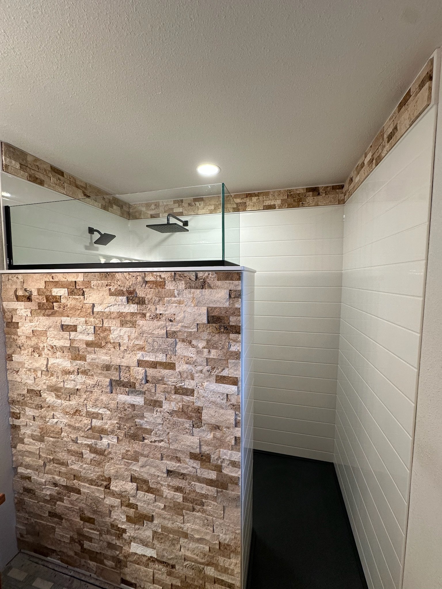 A modern bathroom shower area with a partial glass wall, stone tile accents, white shiplap walls, a ceiling light, and ceiling-mounted rainfall showerheads.