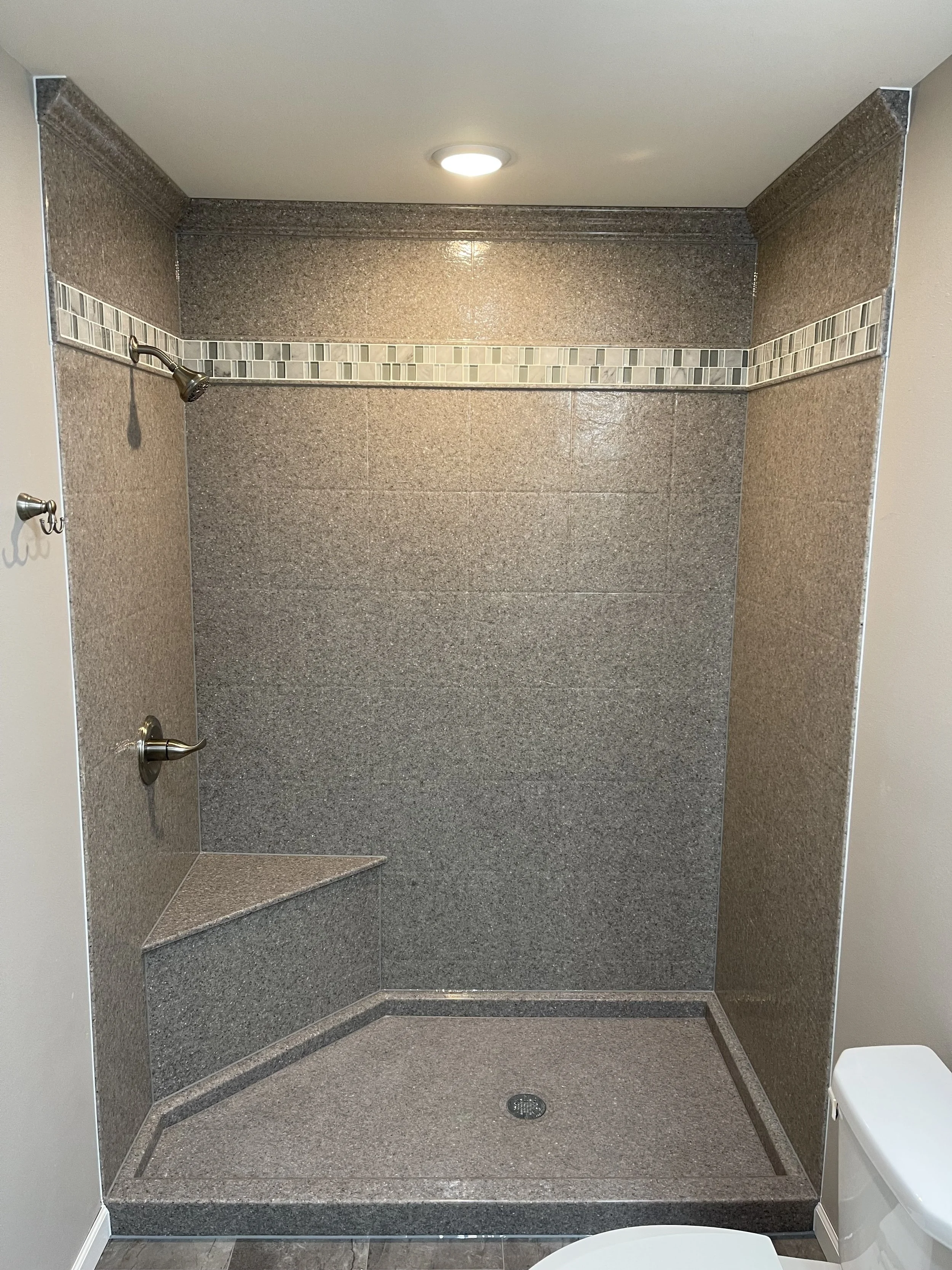 A spacious walk-in shower with brown and beige tiled walls, a decorative tile border, a built-in corner seat, and a ceiling light. Part of a white toilet is visible in the foreground.