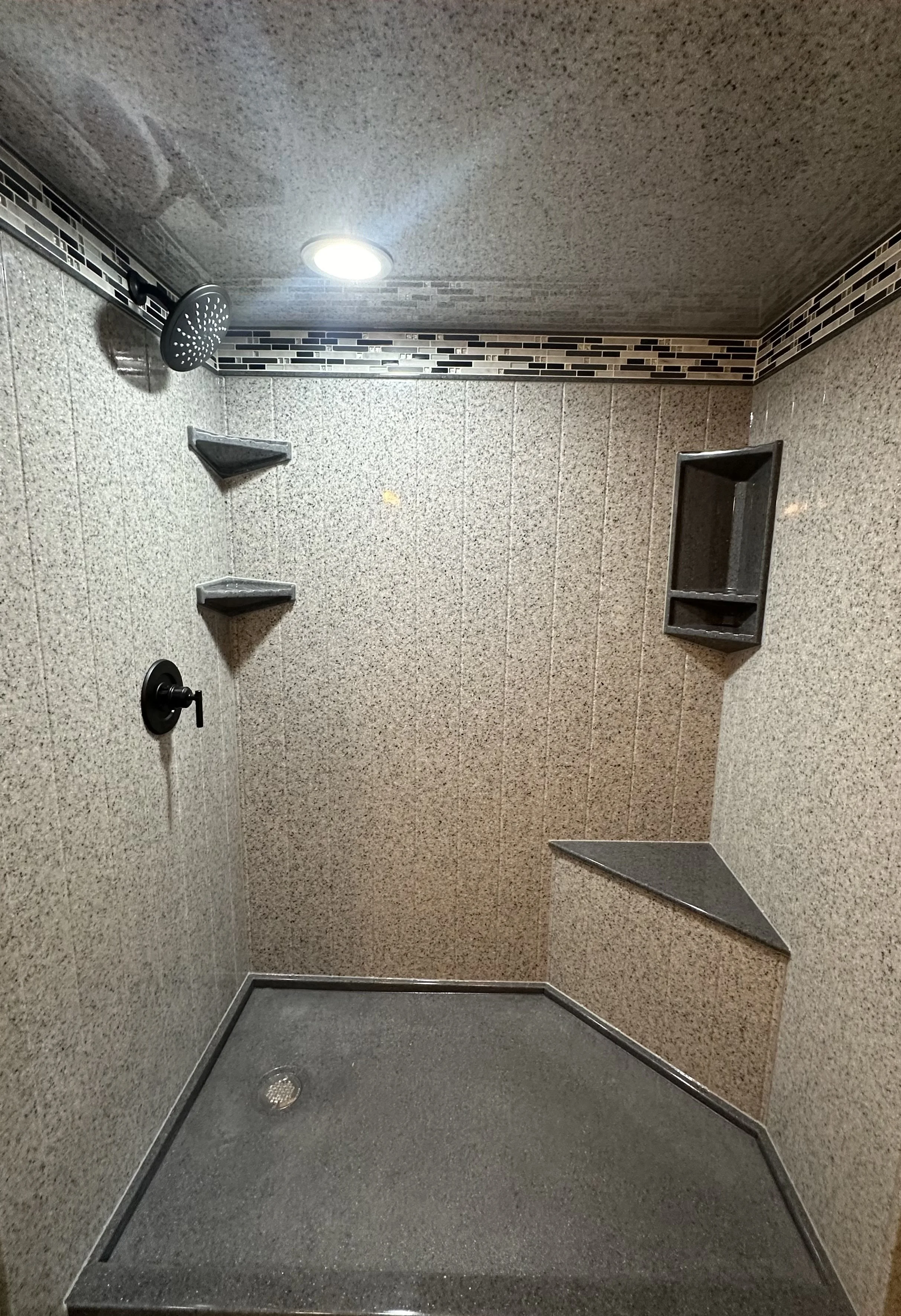 Empty shower stall with textured walls, a showerhead, two small corner shelves, a built-in soap dish, a niche for toiletries, and a textured non-slip floor.