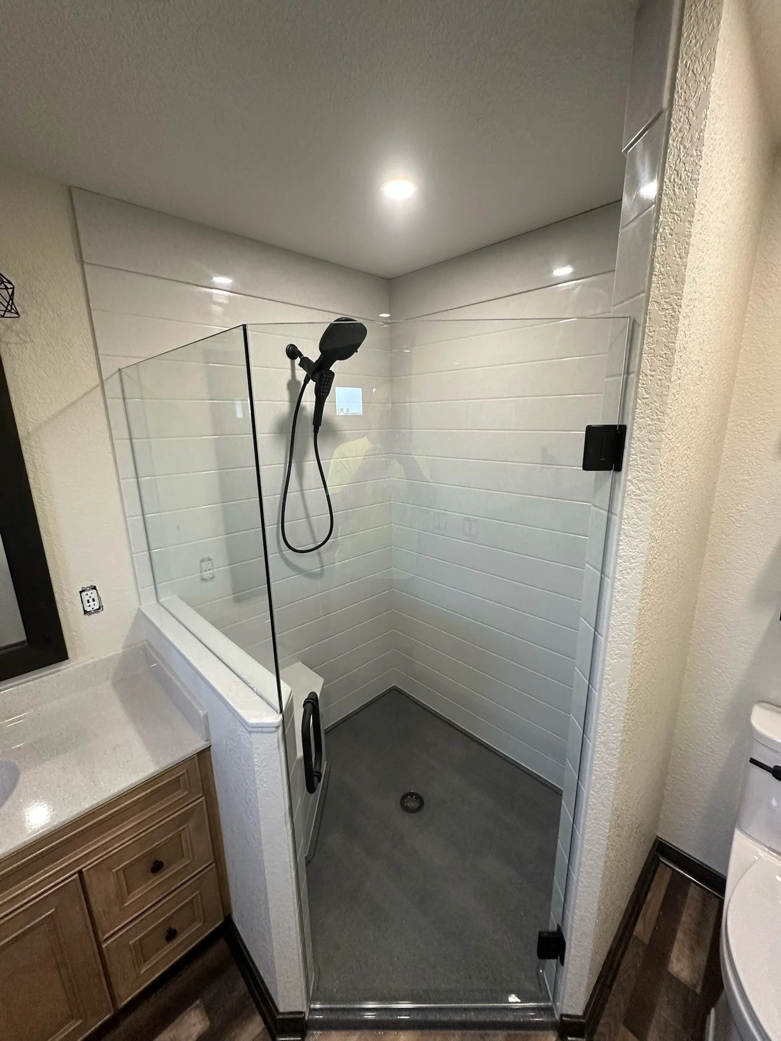 A modern walk-in shower with glass enclosure, white tiled walls, a black handheld showerhead, and a dark gray floor, next to a bathroom vanity and toilet.