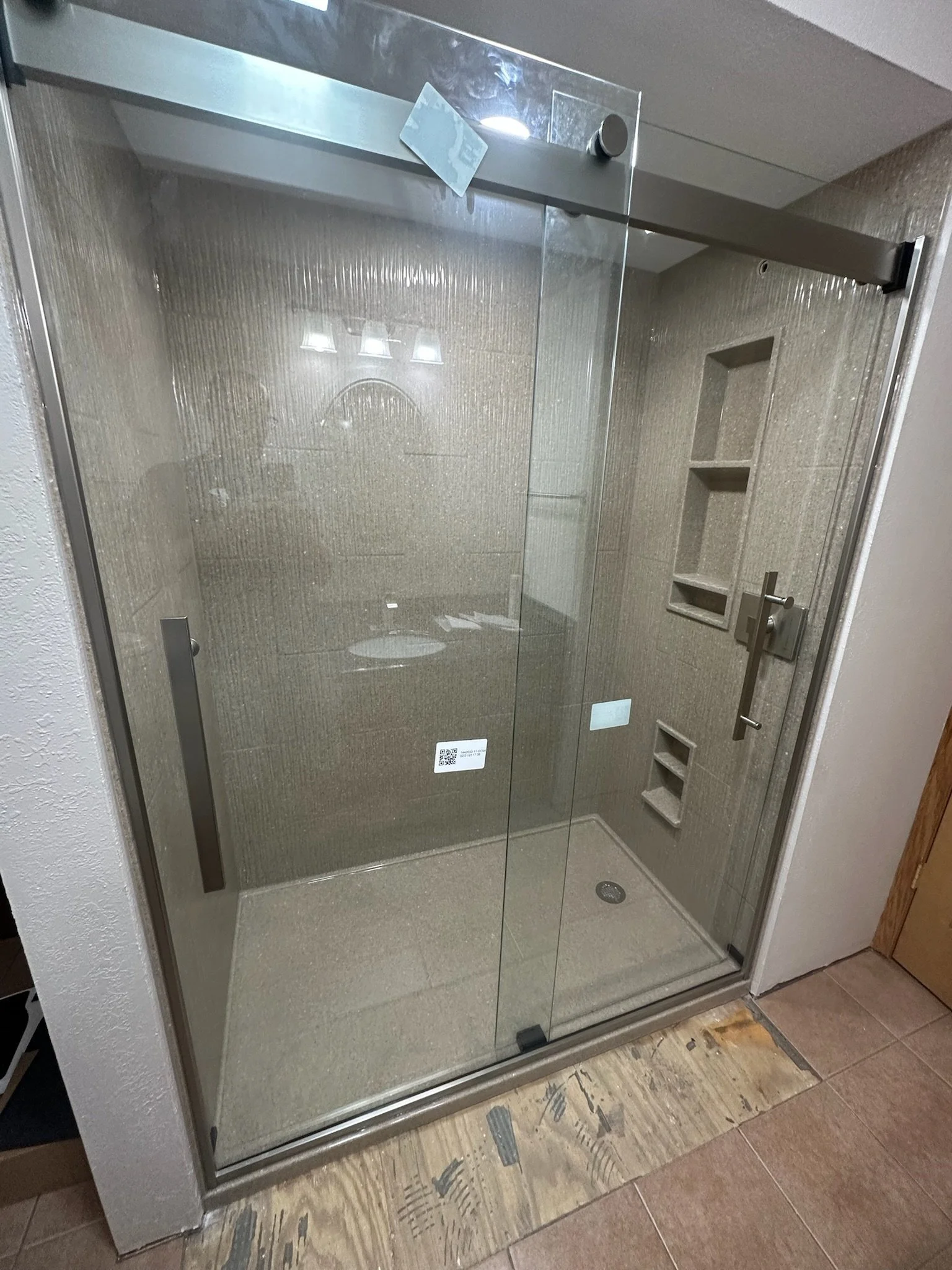 A glass shower enclosure with metal frame, sliding door, and built-in wall niche and shelves. Reflection of lights on the glass, with a tiled interior and a drain at the shower floor.