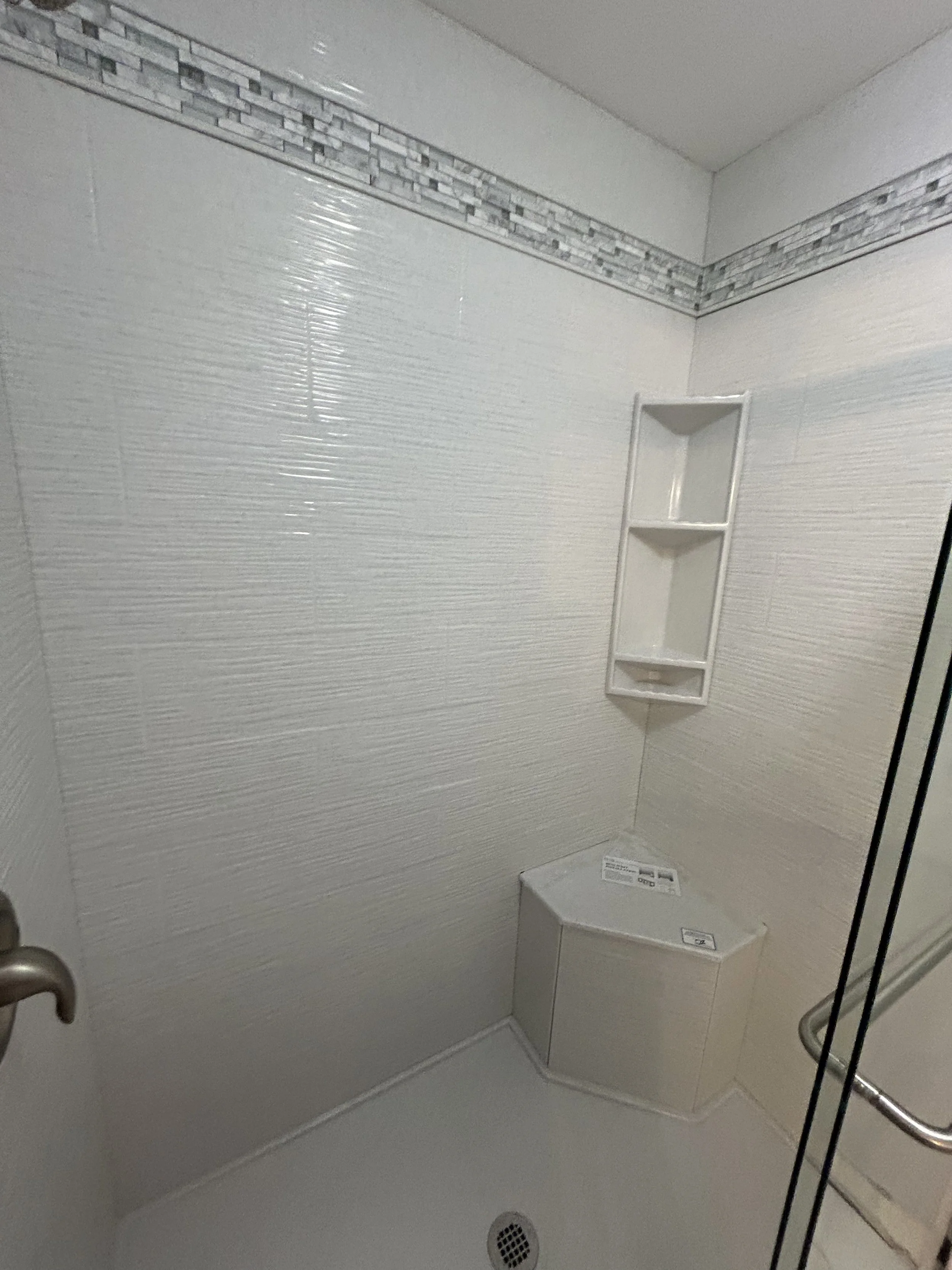 White tiled shower with a built-in corner shelf and a small rectangular bench, with a drain on the floor and a partial view of a glass door and door handle.