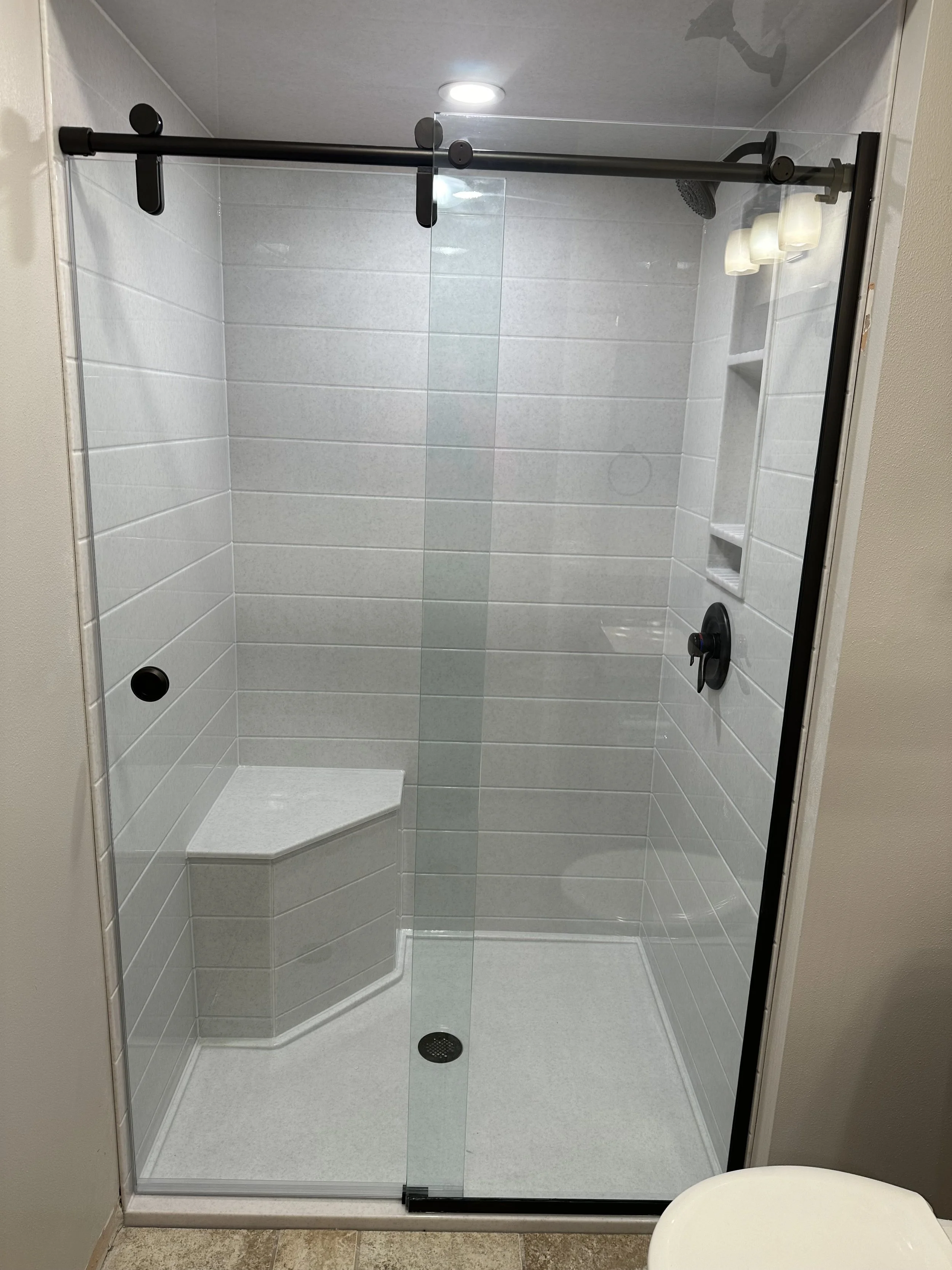 A modern walk-in shower with white tiled walls, black fixtures, a built-in corner seat, sliding glass door, and a decorative niche for toiletries.
