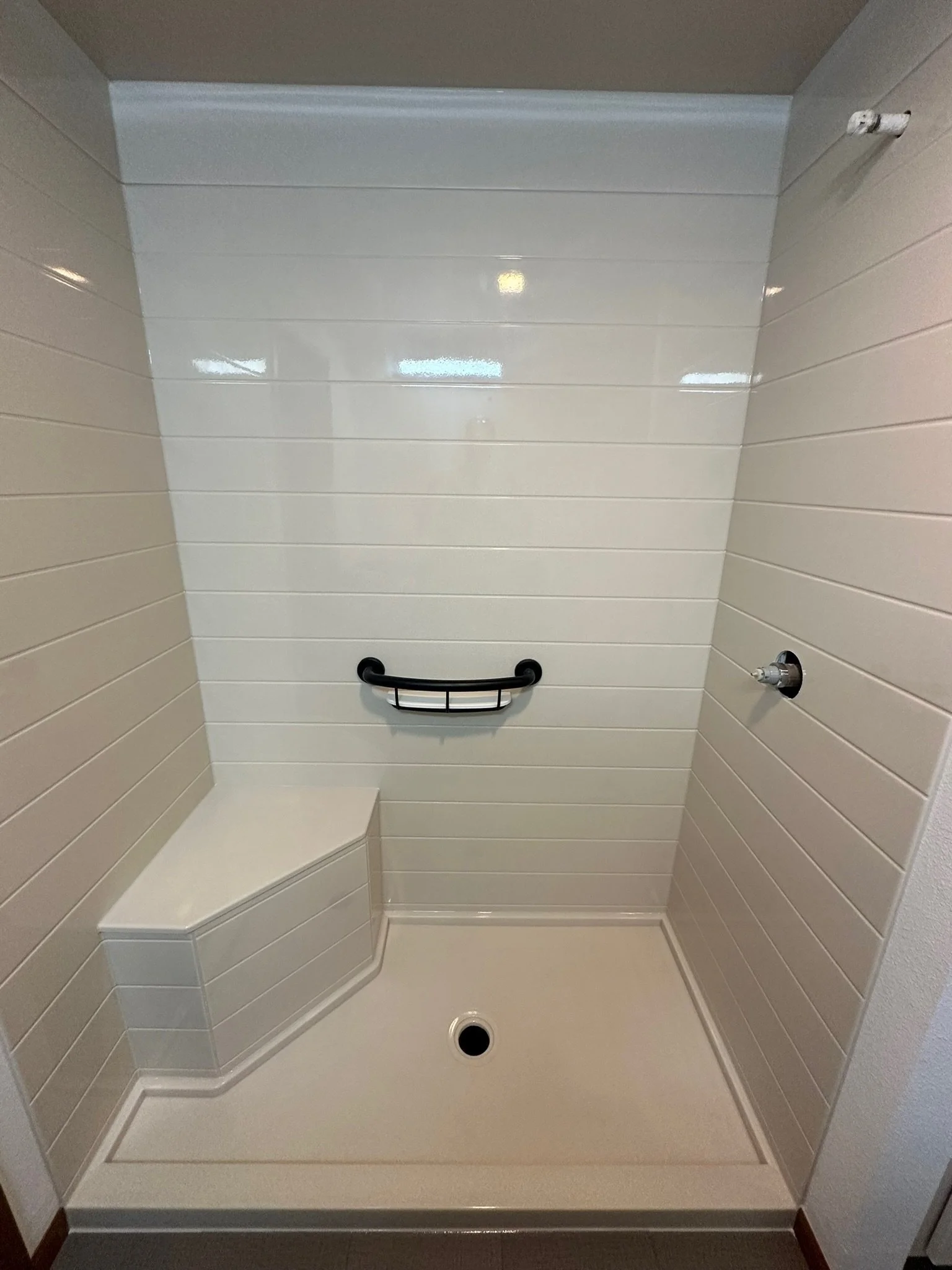 Empty shower stall with cream-colored tiled walls, a black grab bar, a built-in corner seat, and a drain at the bottom.