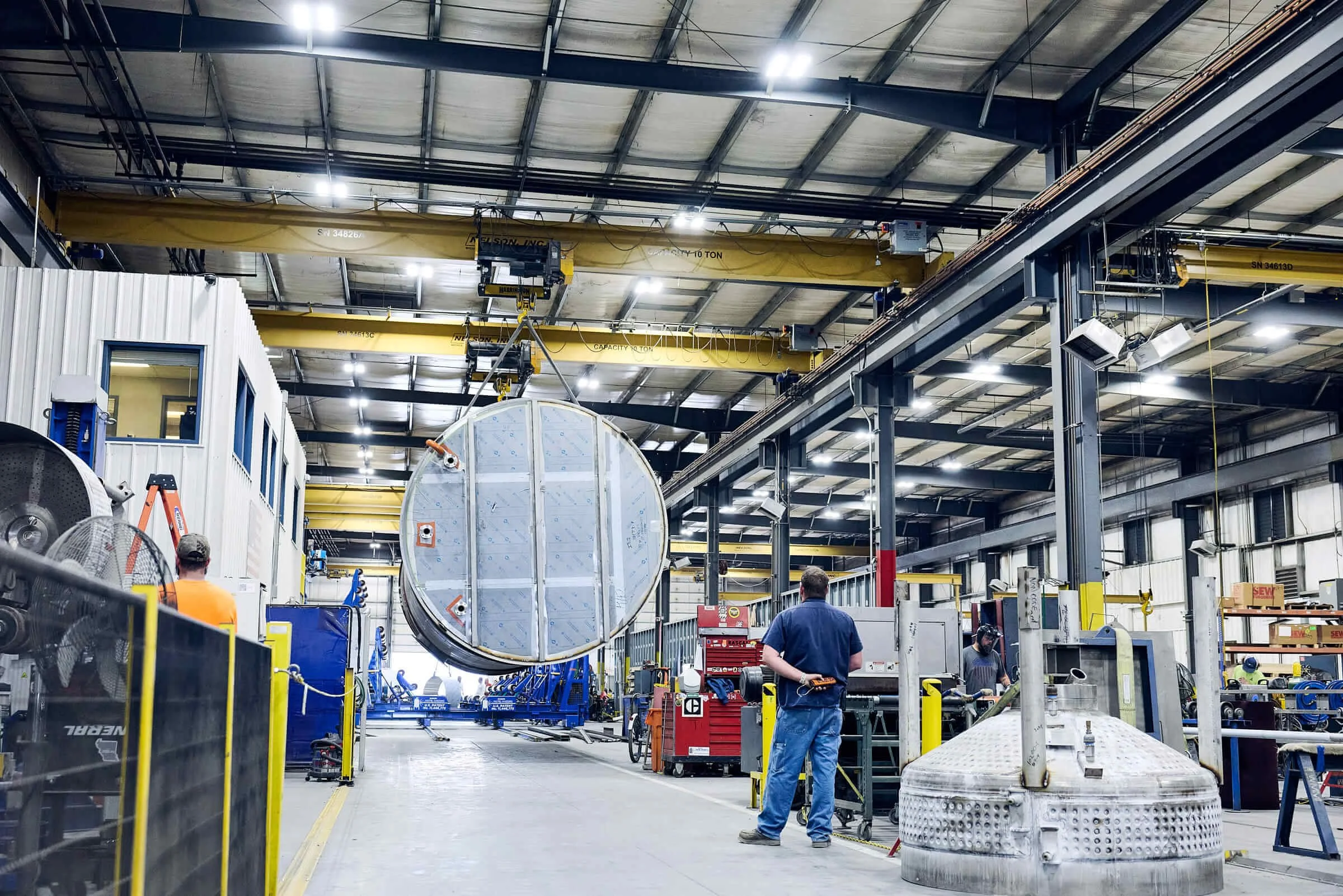Workers in a large industrial factory with high ceilings, yellow overhead cranes, and large machinery, assembling a stainless steel tank.