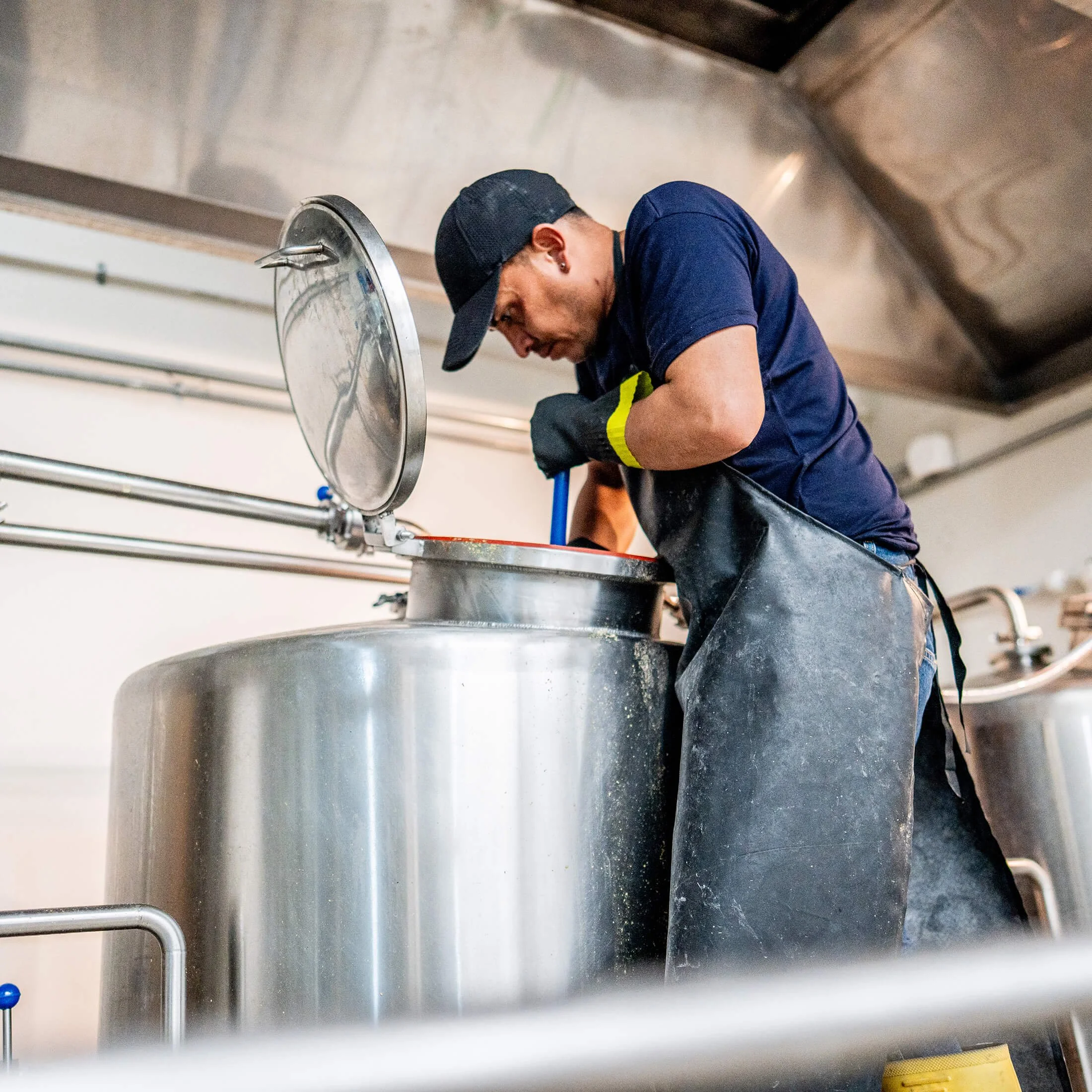 A person wearing a black baseball cap, dark blue shirt, and black gloves is leaning over a large metal tank, possibly working in a commercial kitchen or brewery, with a stainless steel environment in the background.