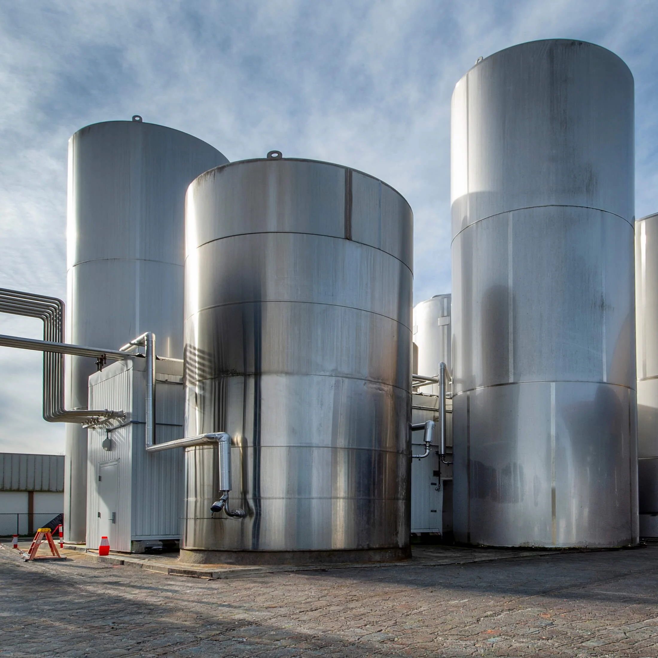 Three large stainless steel storage tank outside near a building, with pipes and pipes near the tanks, and traffic cones and barriers on the cobblestone ground.