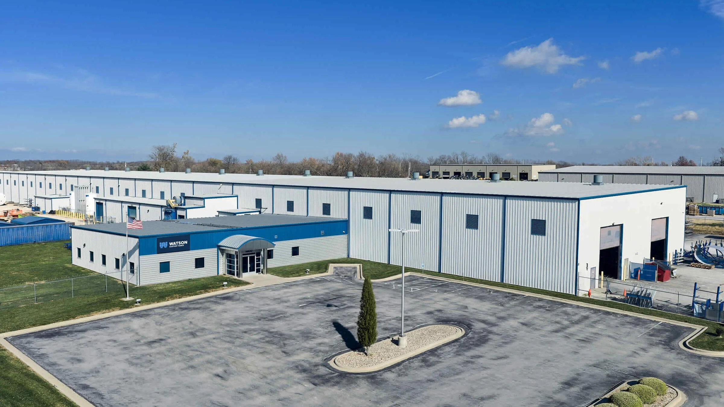 An industrial warehouse complex with a parking lot in the foreground, featuring a blue and gray building with a sign that reads 'Watson.' The large white building has multiple garage doors and a fenced area nearby. The sky is clear with a few clouds.