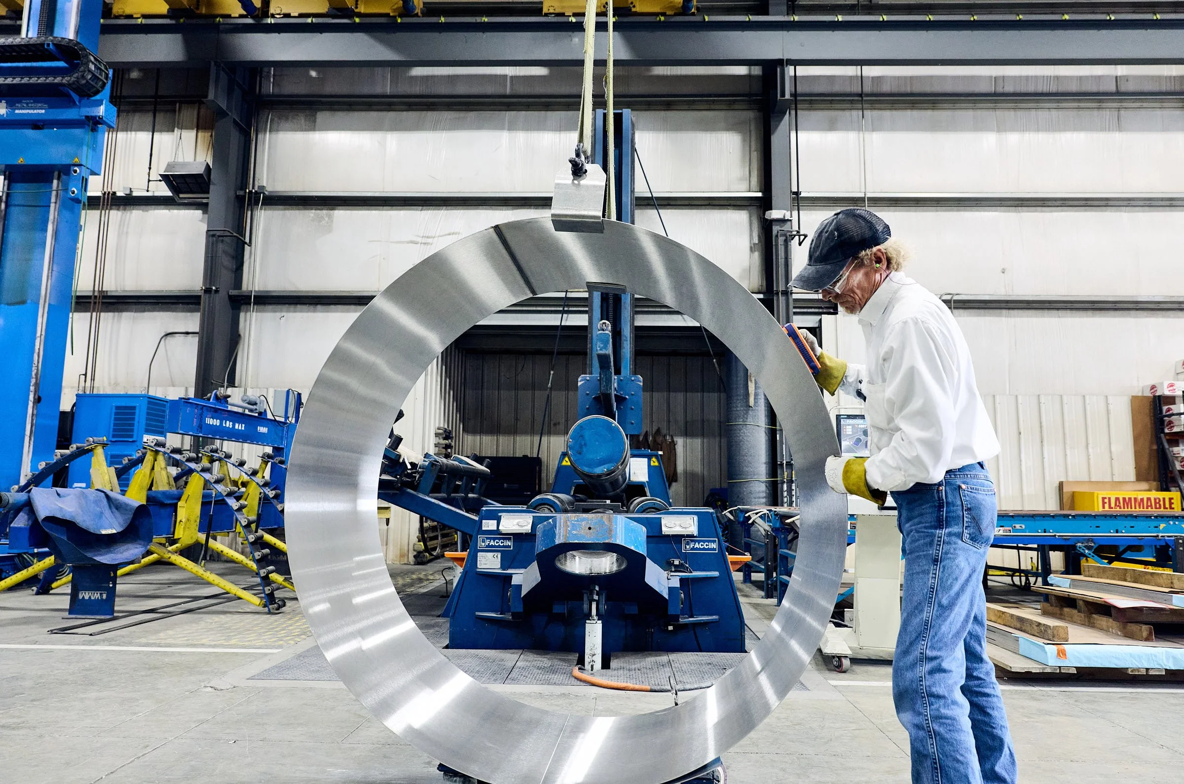 A worker in safety gear inspecting a stainless steel tank component in an industrial setting with machinery and equipment around.