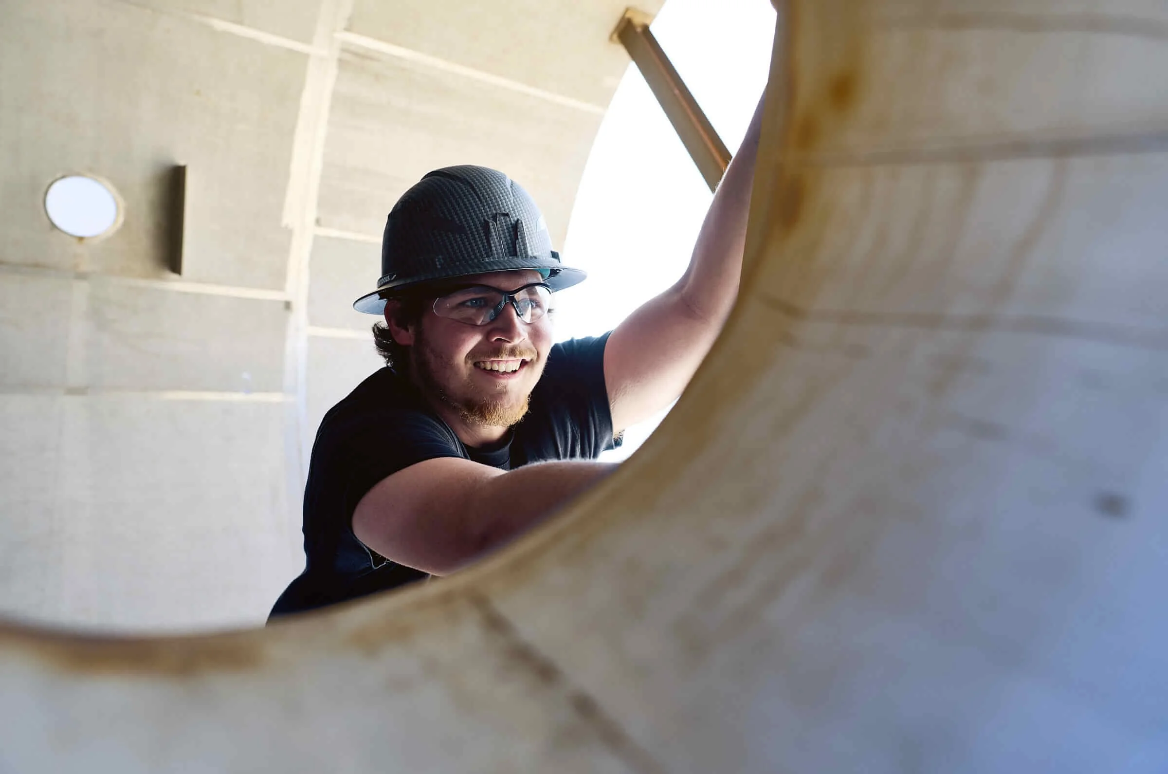 A man wearing a safety helmet and glasses is smiling while working on a surface, seen from underneath.