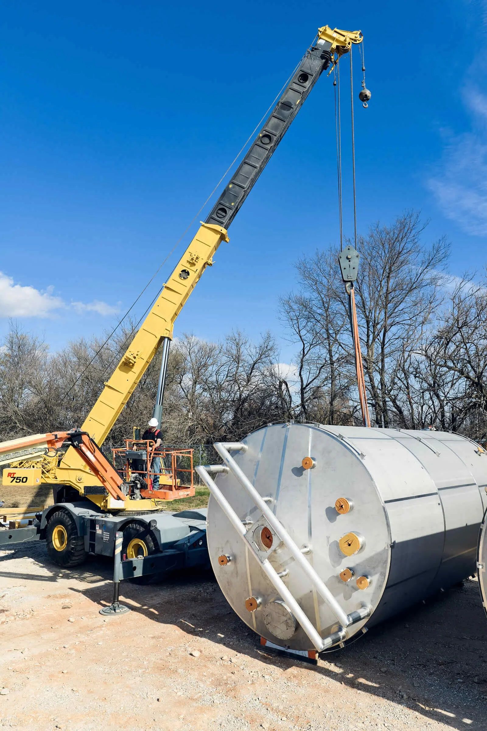 Construction workers using a crane to lift a large stainless steel tank with orange bolts in an outdoor setting with trees and a blue sky.