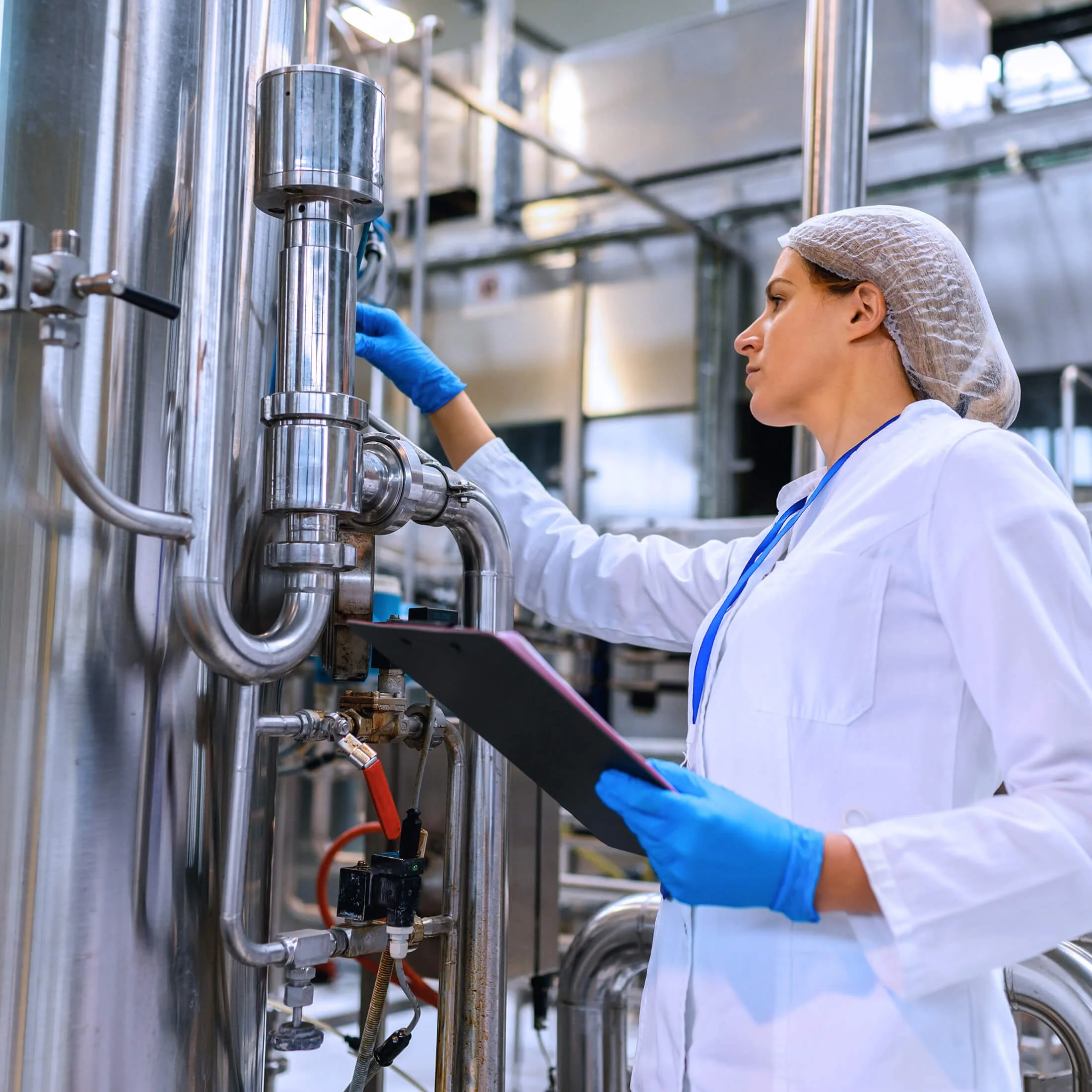 A scientist or technician wearing a white lab coat, hairnet, and blue gloves working with stainless steel pipes and machinery in a laboratory or manufacturing facility.