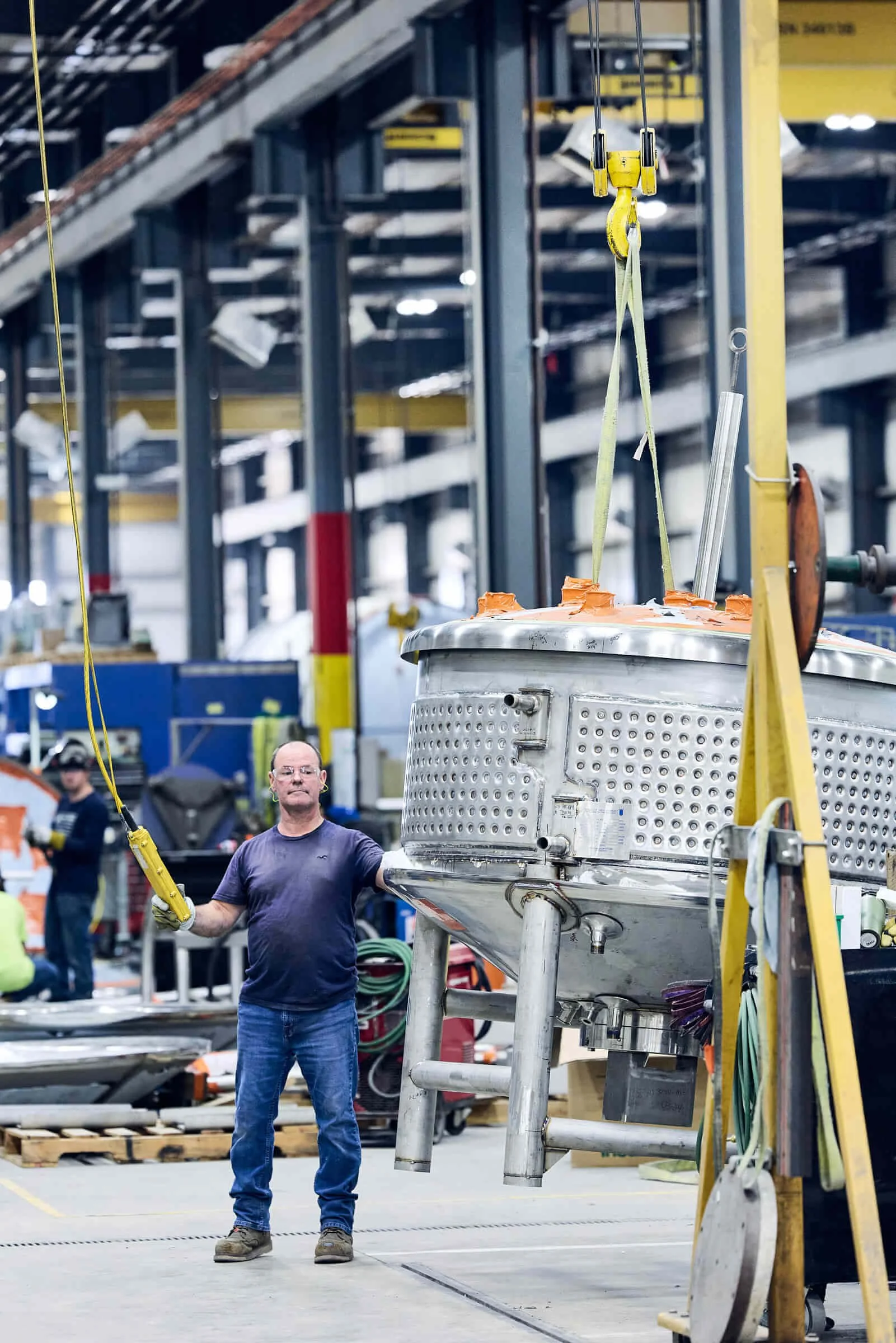 A person in a blue shirt and jeans holding a yellow tool or device, standing in front of a stainless steel tank inside a manufacturing facility. The background shows a spacious, organized workspace with various equipment and workers.