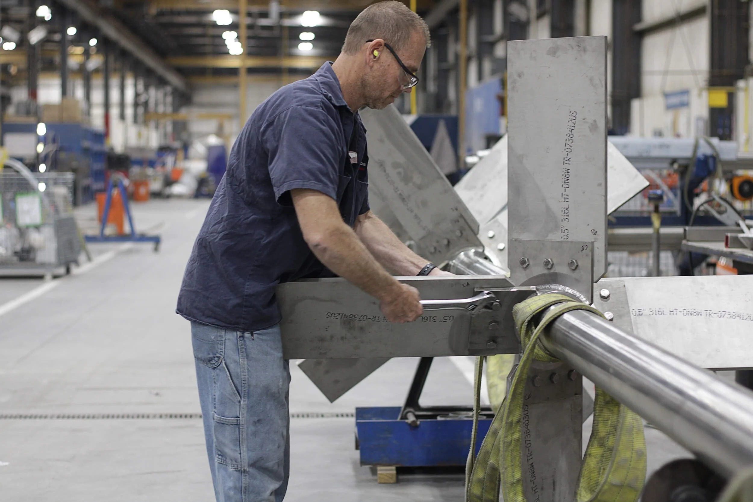A man in safety glasses and yellow ear protection works on a tank impeller in a factory setting. The background features shelves, tools, and equipment.