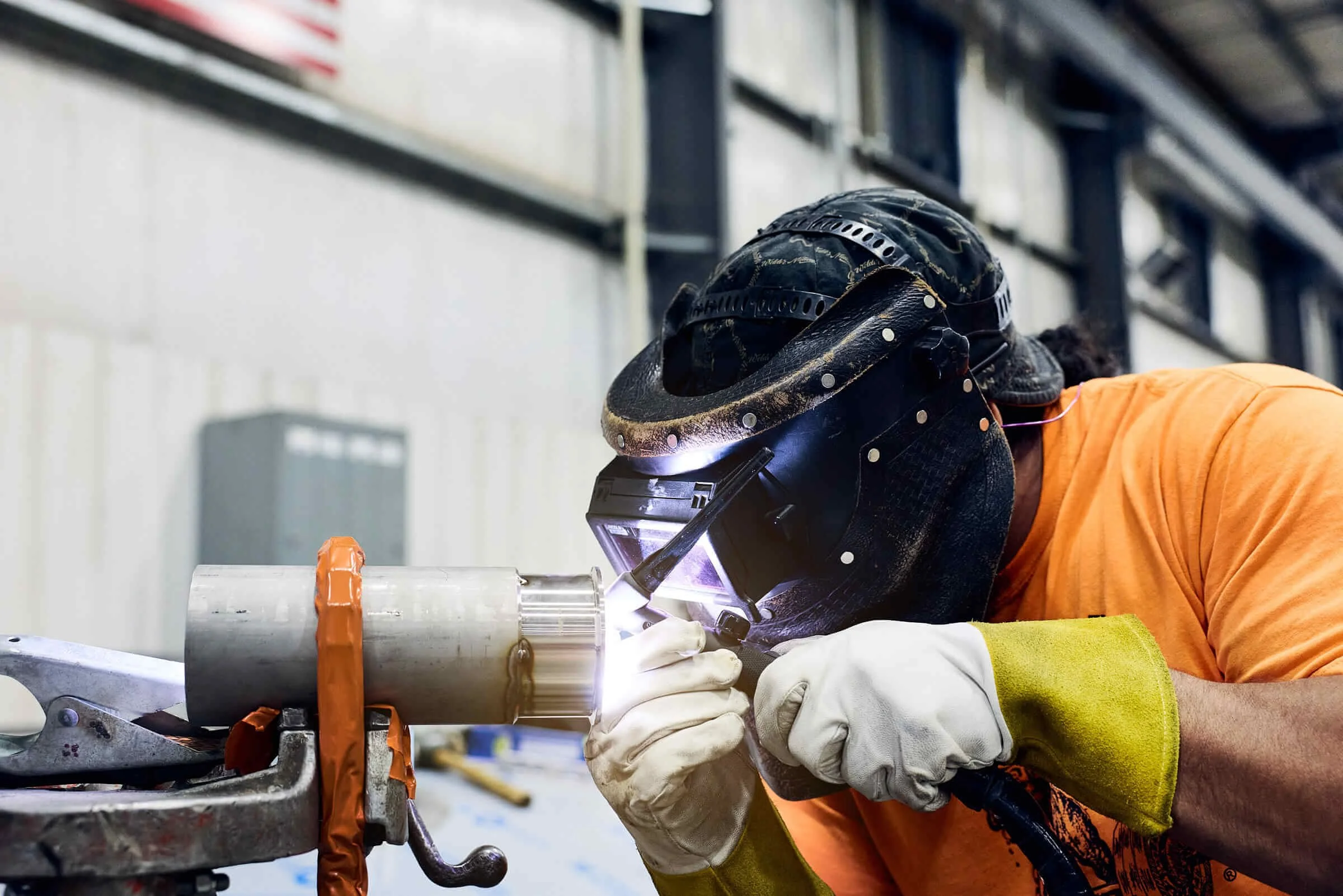 Worker welding a metal pipe in an industrial setting, wearing protective gear including gloves, helmet, and orange safety clothing.