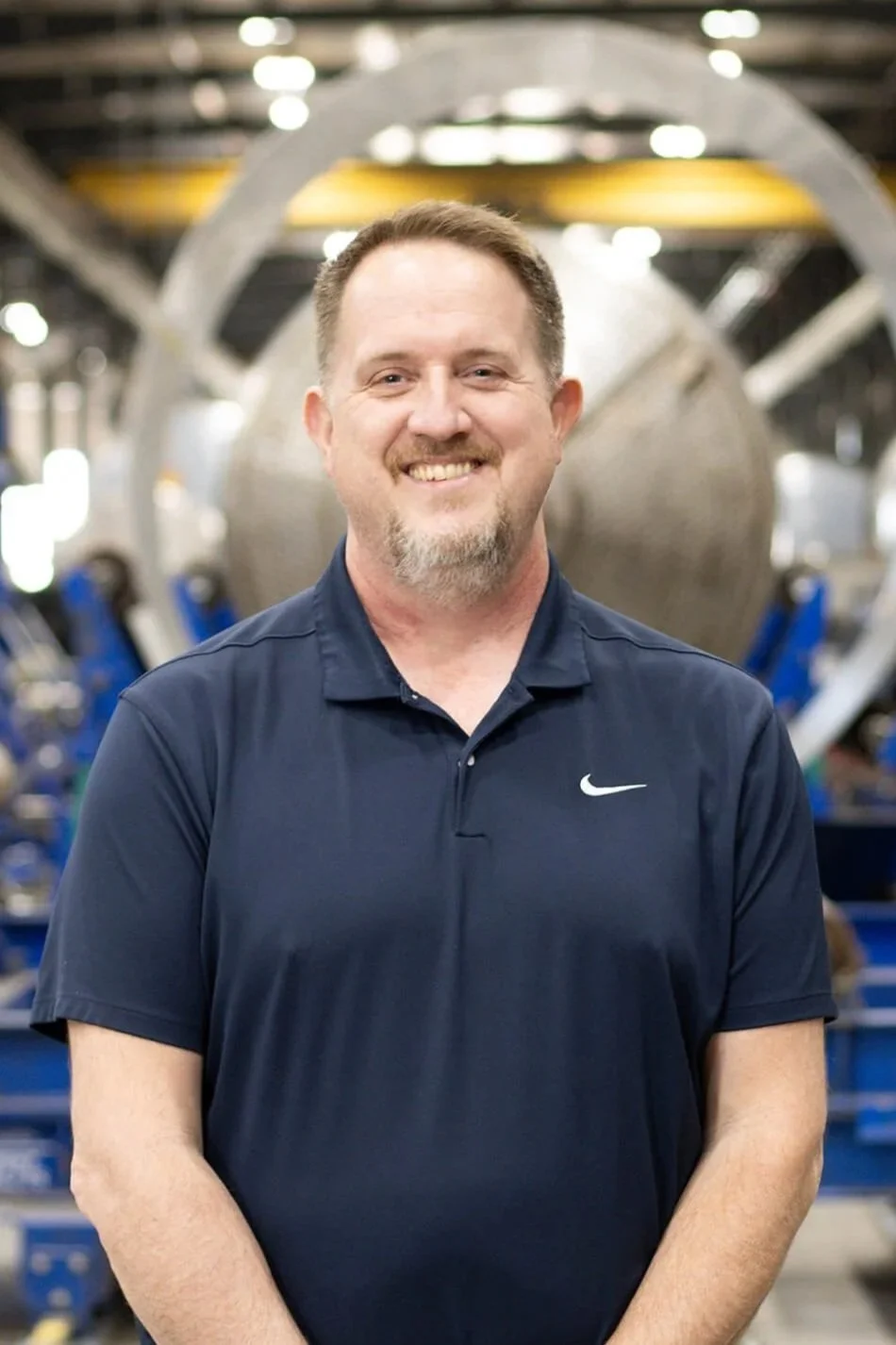 A smiling man with short hair and a beard wearing a navy blue Nike polo shirt, standing inside a large industrial facility or factory with machinery in the background.