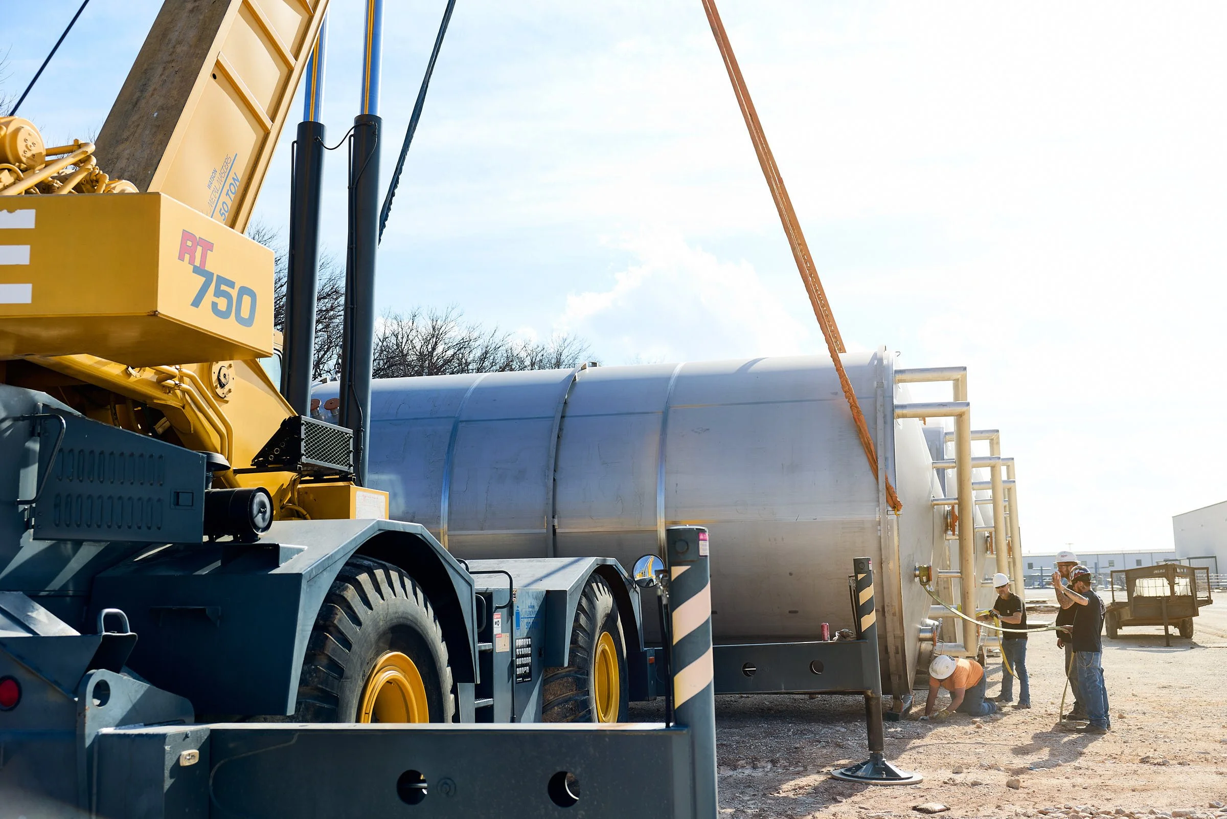 A construction site with a large yellow crane lifting a stainless steel tank. Several workers wearing helmets are working near the tank, and the sky is blue with some clouds.