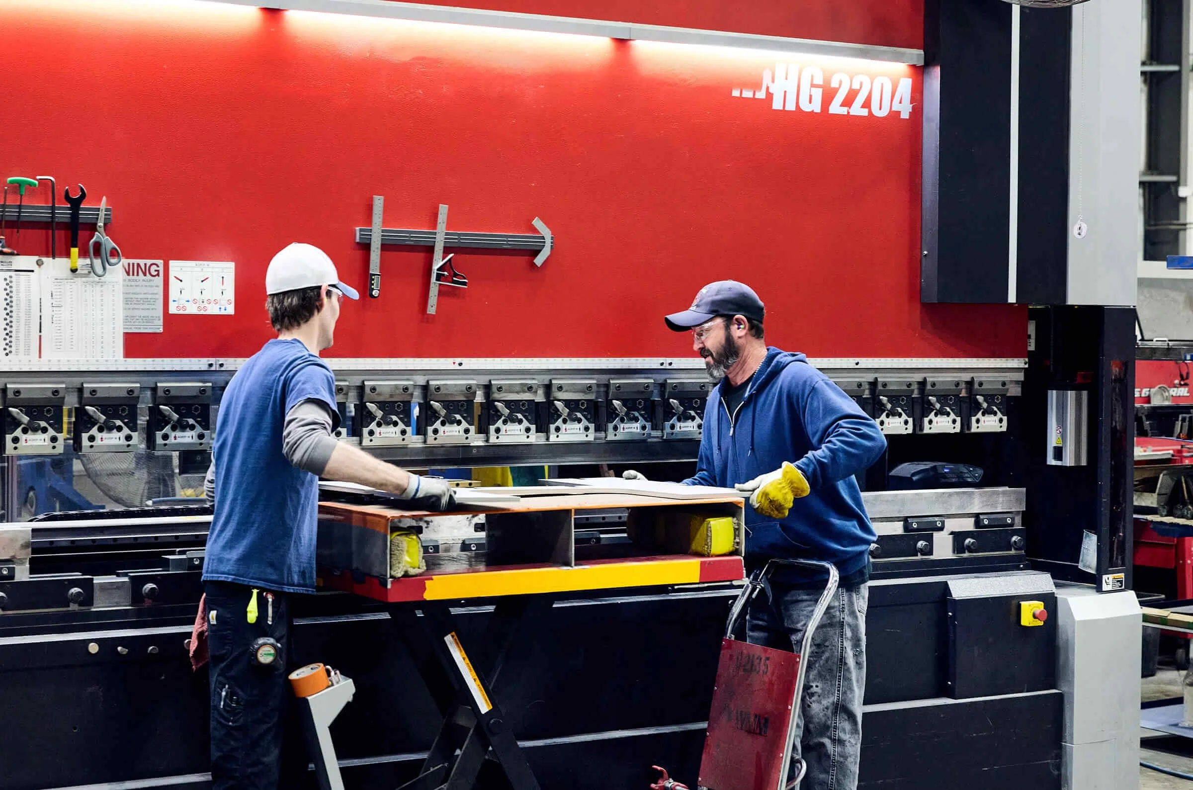 Two workers wearing safety gear and gloves working on a large piece of equipment in an industrial setting with a red wall in the background.
