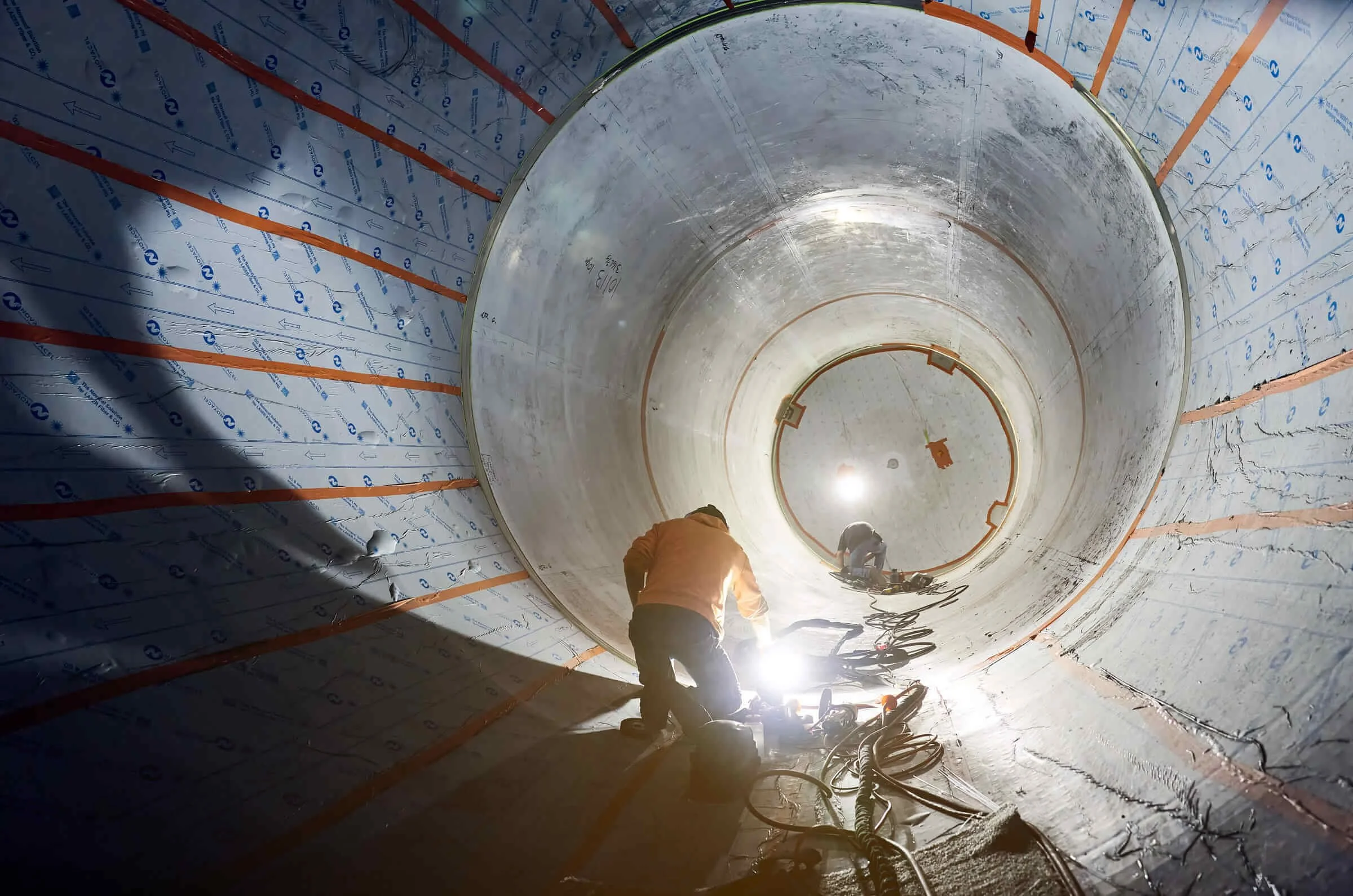Workers working inside a large stainless steel tank, with insulation and orange safety markings.