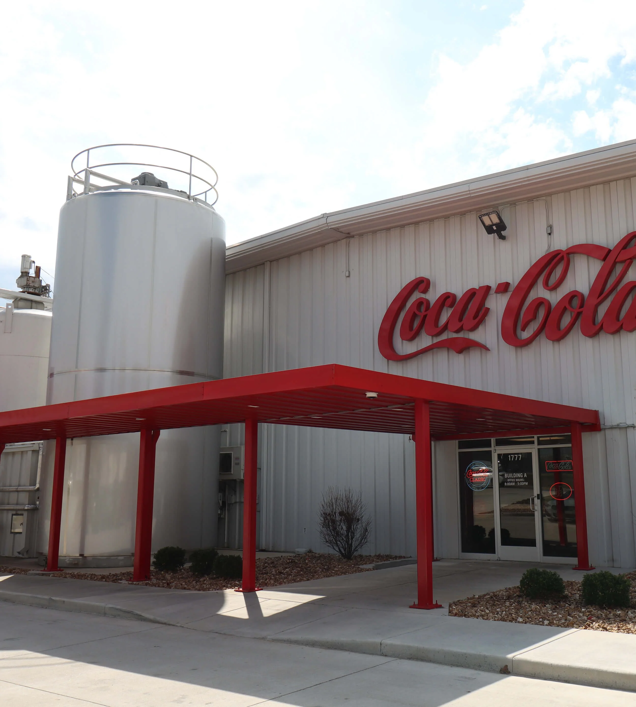 Exterior of a building with the Coca-Cola logo visible, featuring a large white gas storage tank and a red metal canopy, with a small bush and landscaping in front.