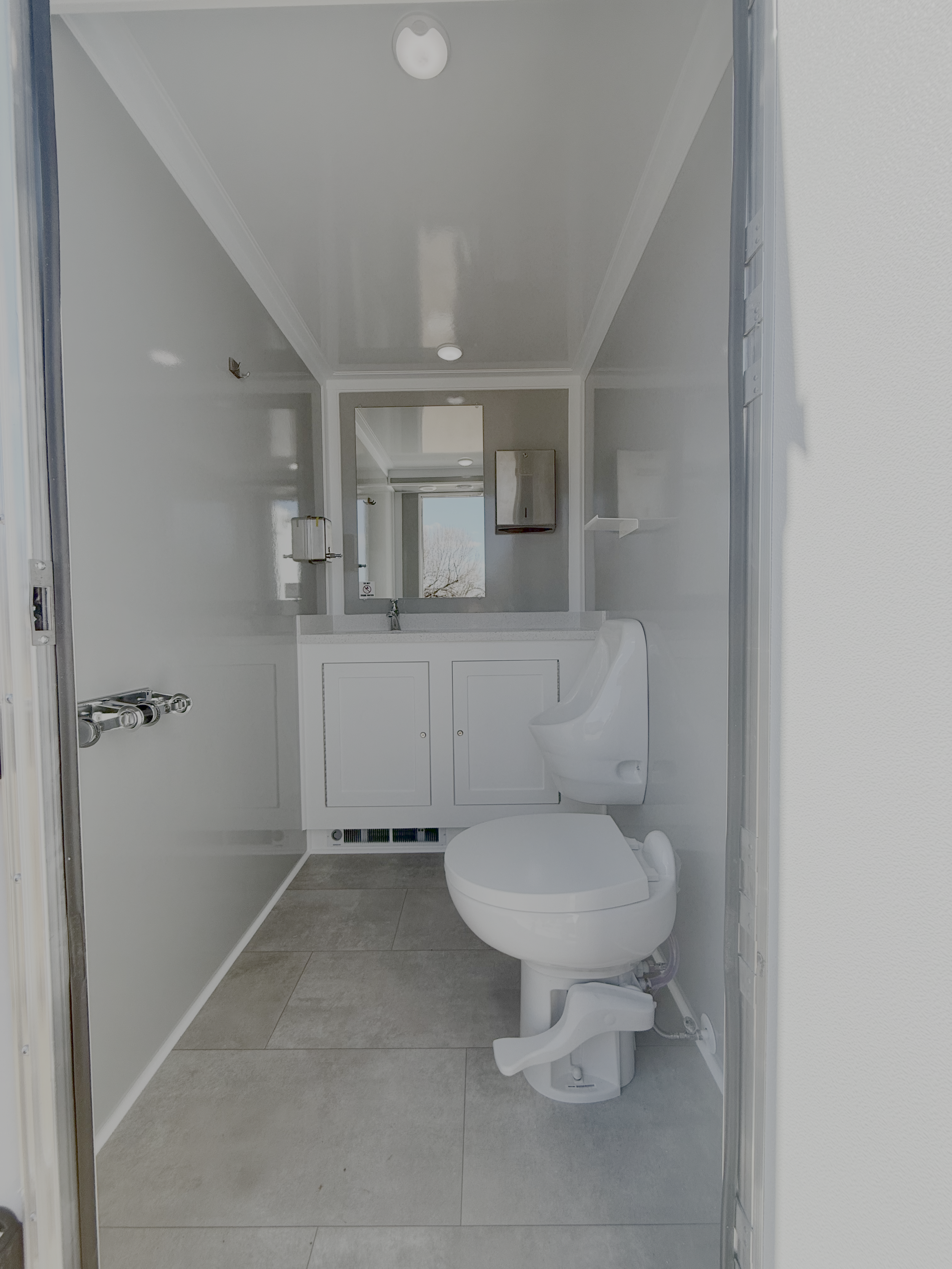 Luxury restroom trailer with a white toilet, a white vanity with a mirror above it, a wall-mounted soap dispenser, and a small shelf. Light gray tiled floor and gray walls.