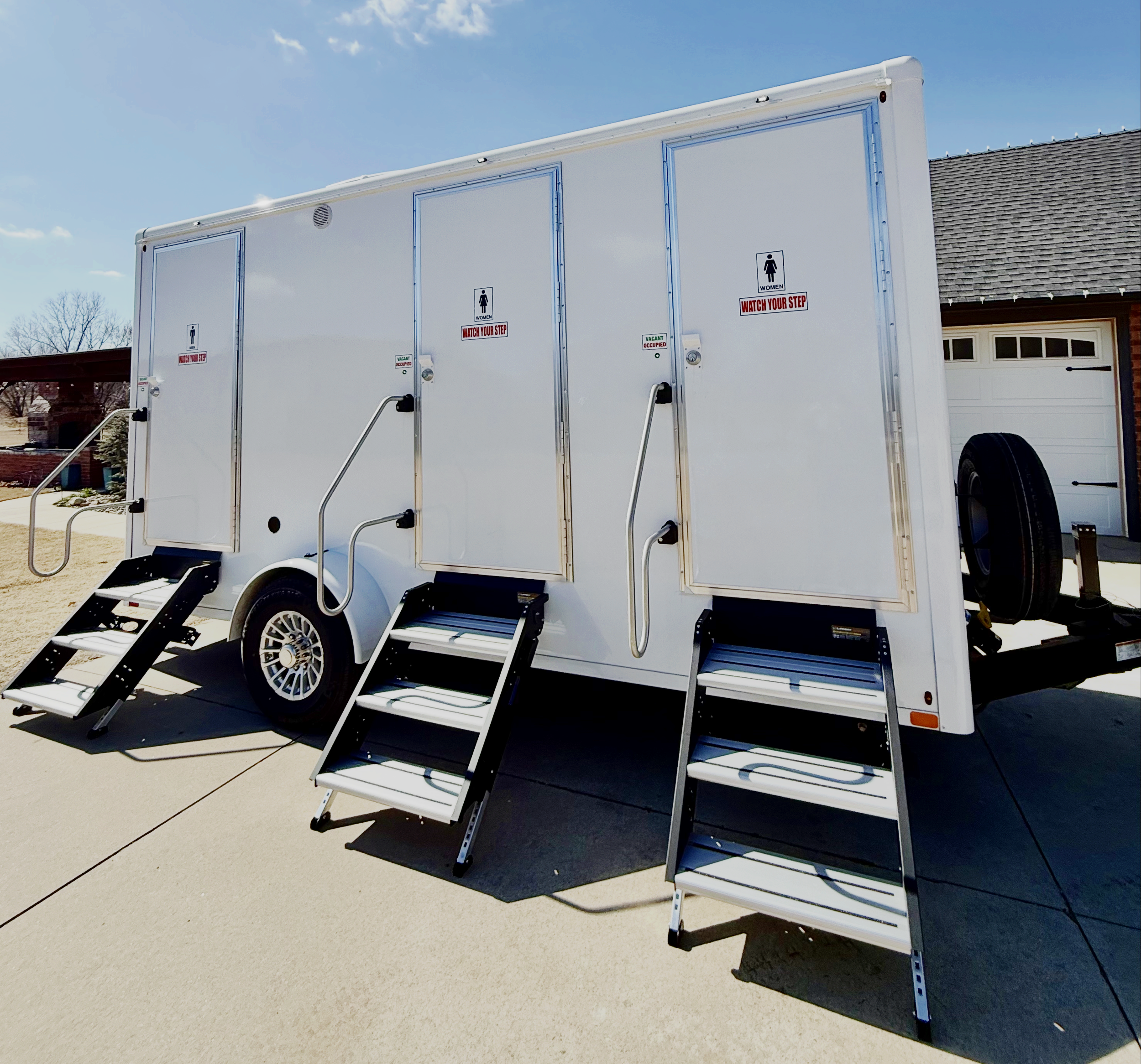 Luxury mobile outdoor restroom trailer with three doors, step stairs, and handrails, parked on a concrete driveway in a suburban neighborhood.