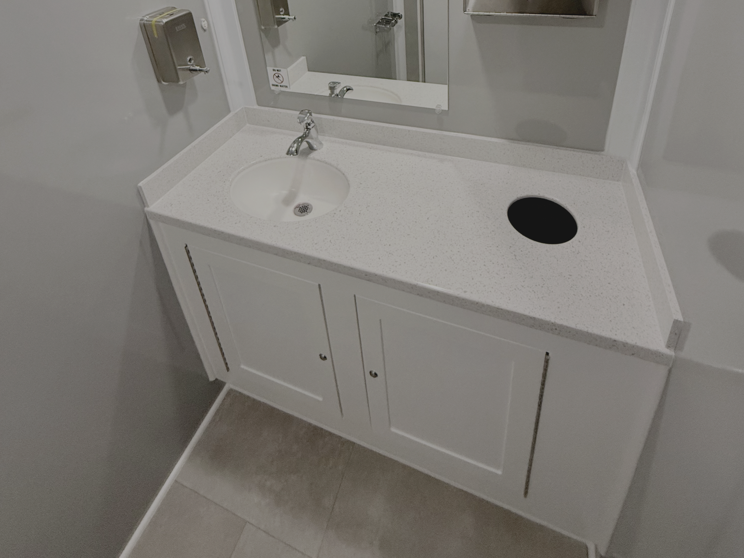 Luxury restroom trailer with white bathroom sink vanity with a small round sink, a faucet, a mirror above, a paper towel dispenser, and a circular opening for trash on the countertop.