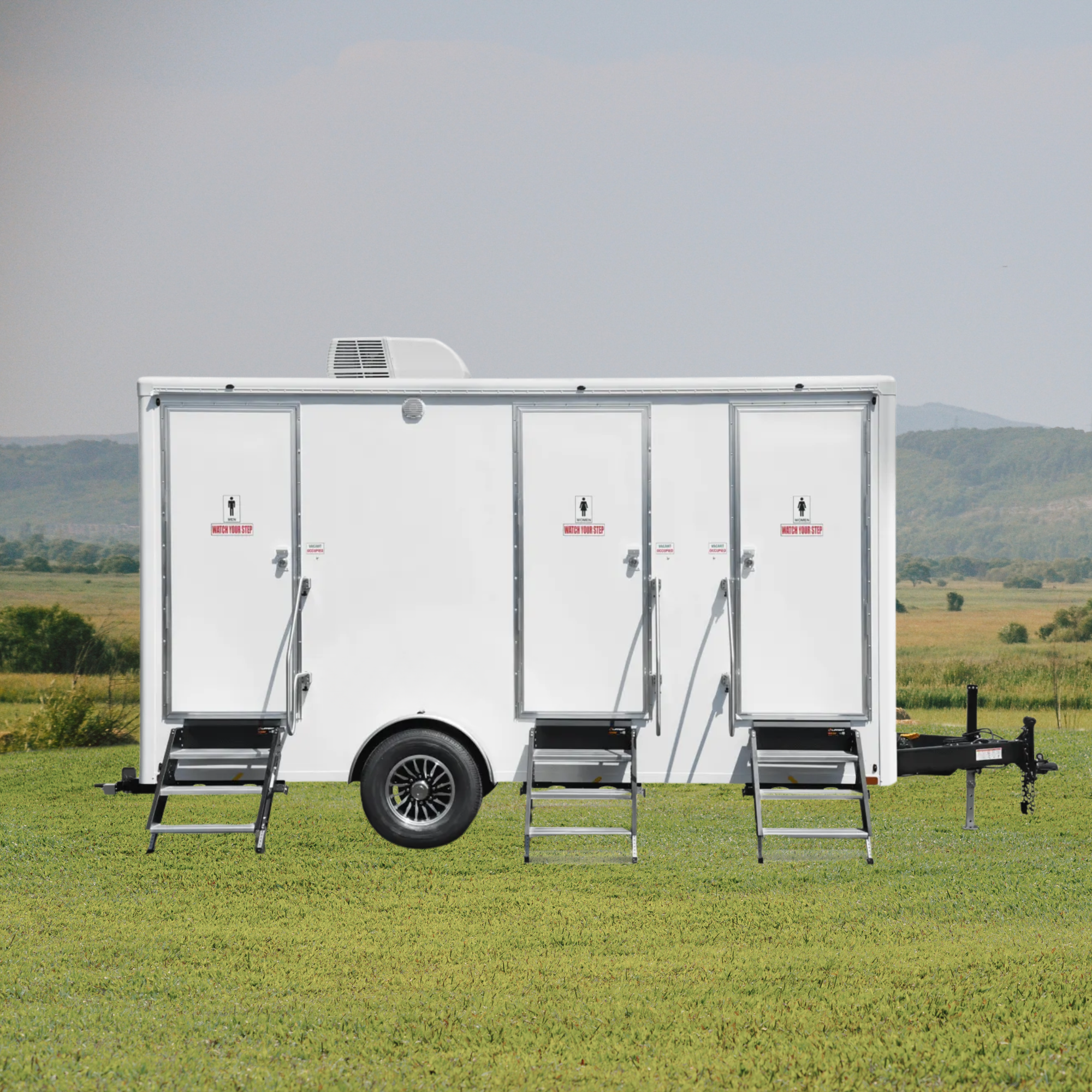 A white luxury restroom trailer with three doors, each with steps and signs indicating unisex facilities, set on a grassy field with distant hills under a cloudy sky.
