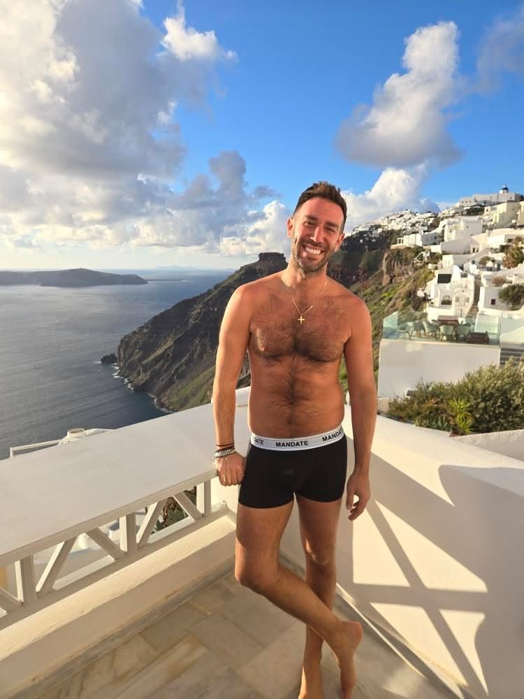 Smiling man in the black Mandate Signature Boxer Brief on a whitewashed balcony above the sea