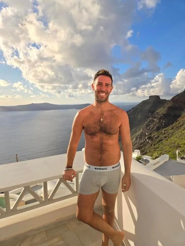 A man standing on a balcony overlooking a body of water and mountainous terrain, wearing gray boxer briefs and smiling.