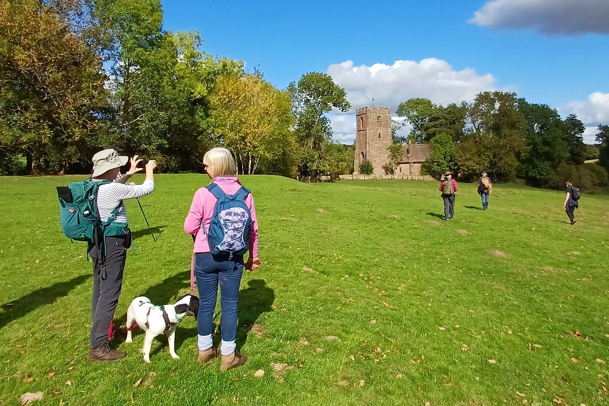 Group of hikers, including two women and a dog, walking on a grassy hill in front of a historic stone castle surrounded by trees on a sunny day.