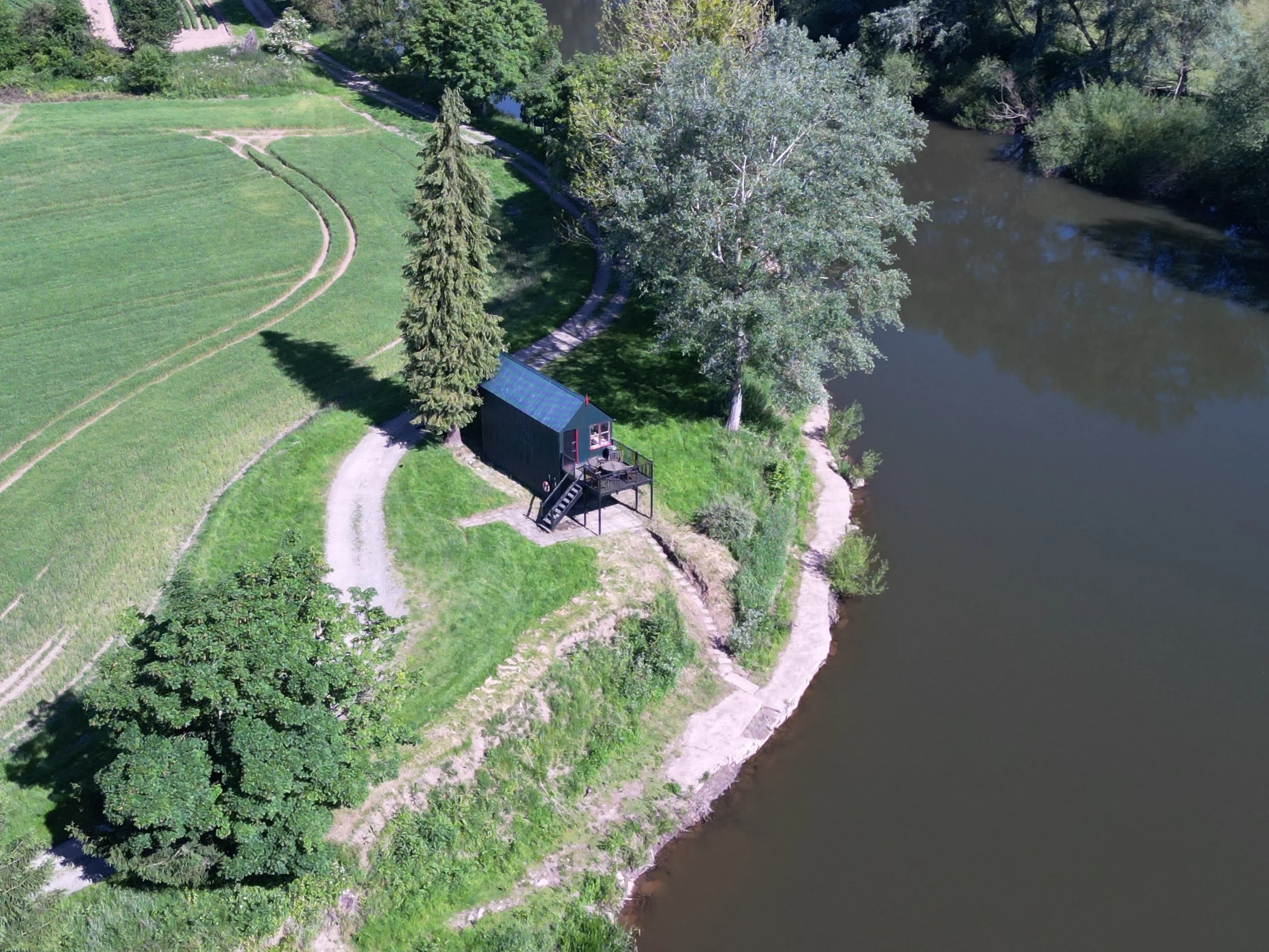 Aerial view of a small dark-colored cabin with stairs on a grassy area beside a river, surrounded by trees and open land.