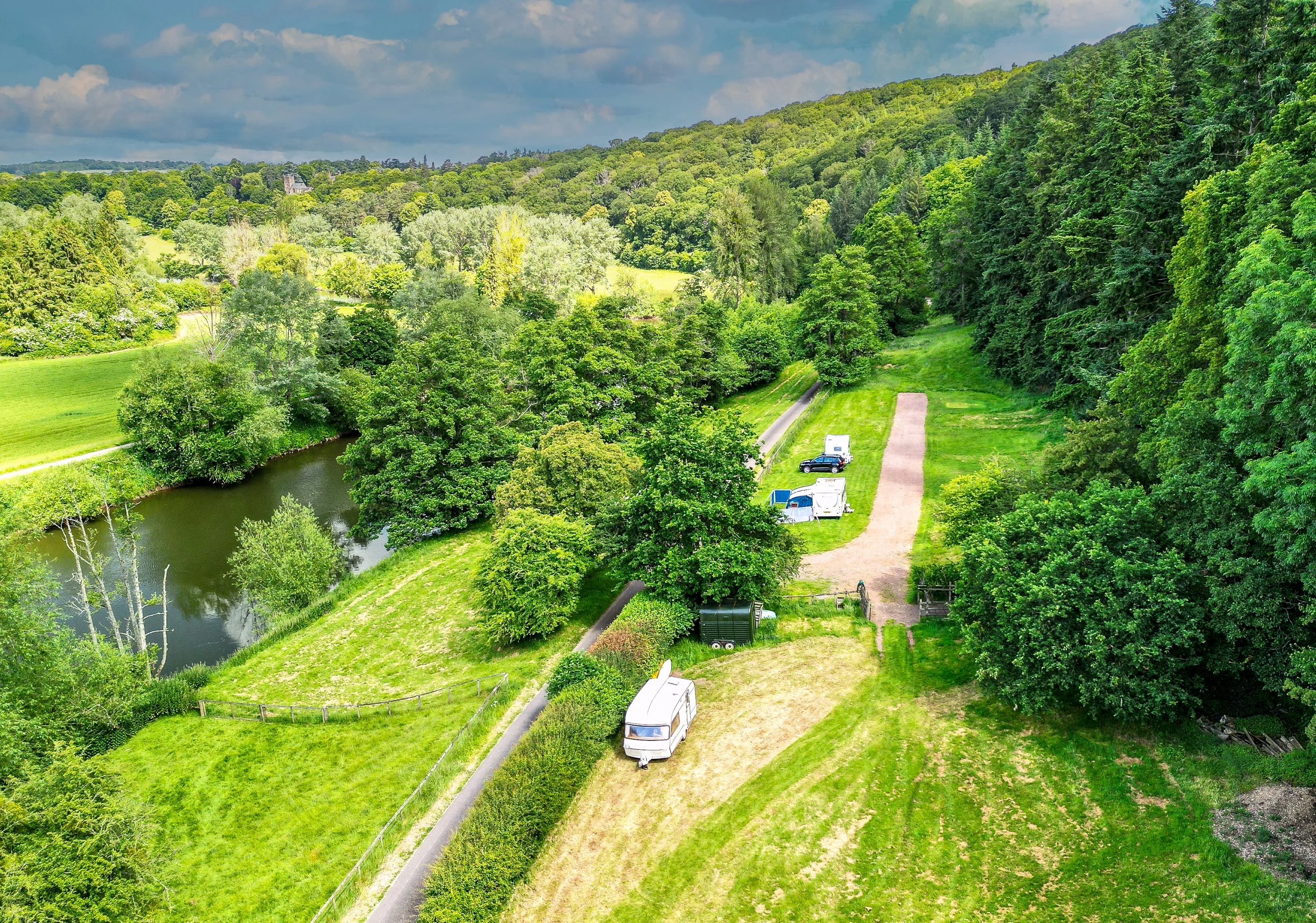 A lush green countryside with a river, trees, grassy areas, and several parked vehicles including a camper, a truck, and a car along gravel and dirt paths.