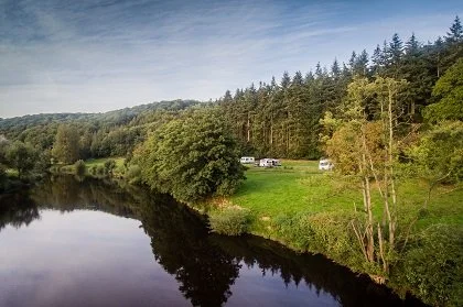 A river reflecting a green landscape with trees and camping trailers in the background.