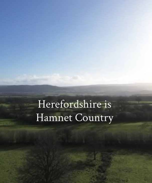 A scenic view of a rural landscape with fields and trees, under a blue sky with clouds, with overlaid text saying "Herefordshire is Hamnet Country."