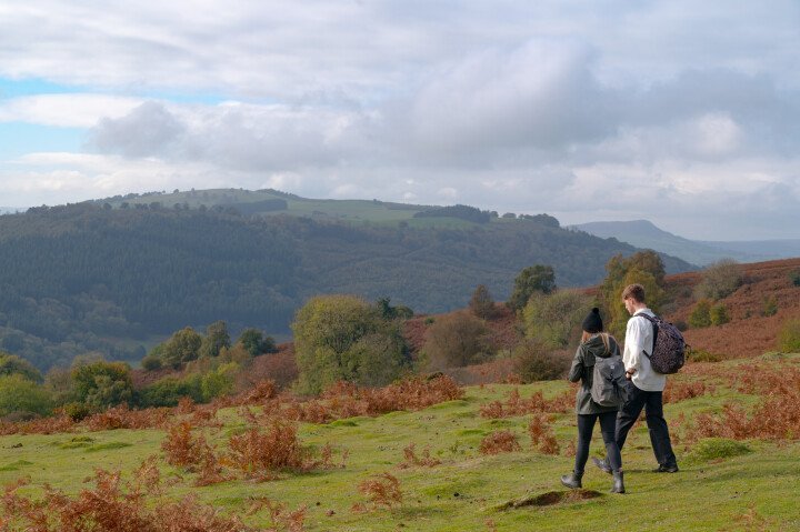 Two people walking on a grassy hillside with rolling hills and a cloudy sky in the background.