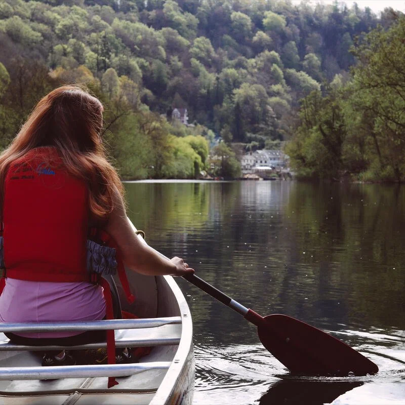 Person wearing a red life jacket paddling a boat on a calm river surrounded by green trees and hills.