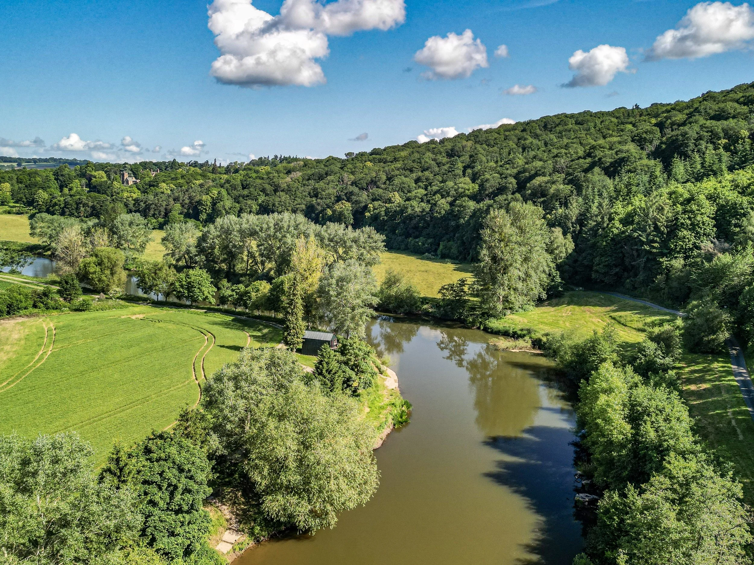 Aerial view of a river winding through a green landscape with trees and fields under a partly cloudy sky.