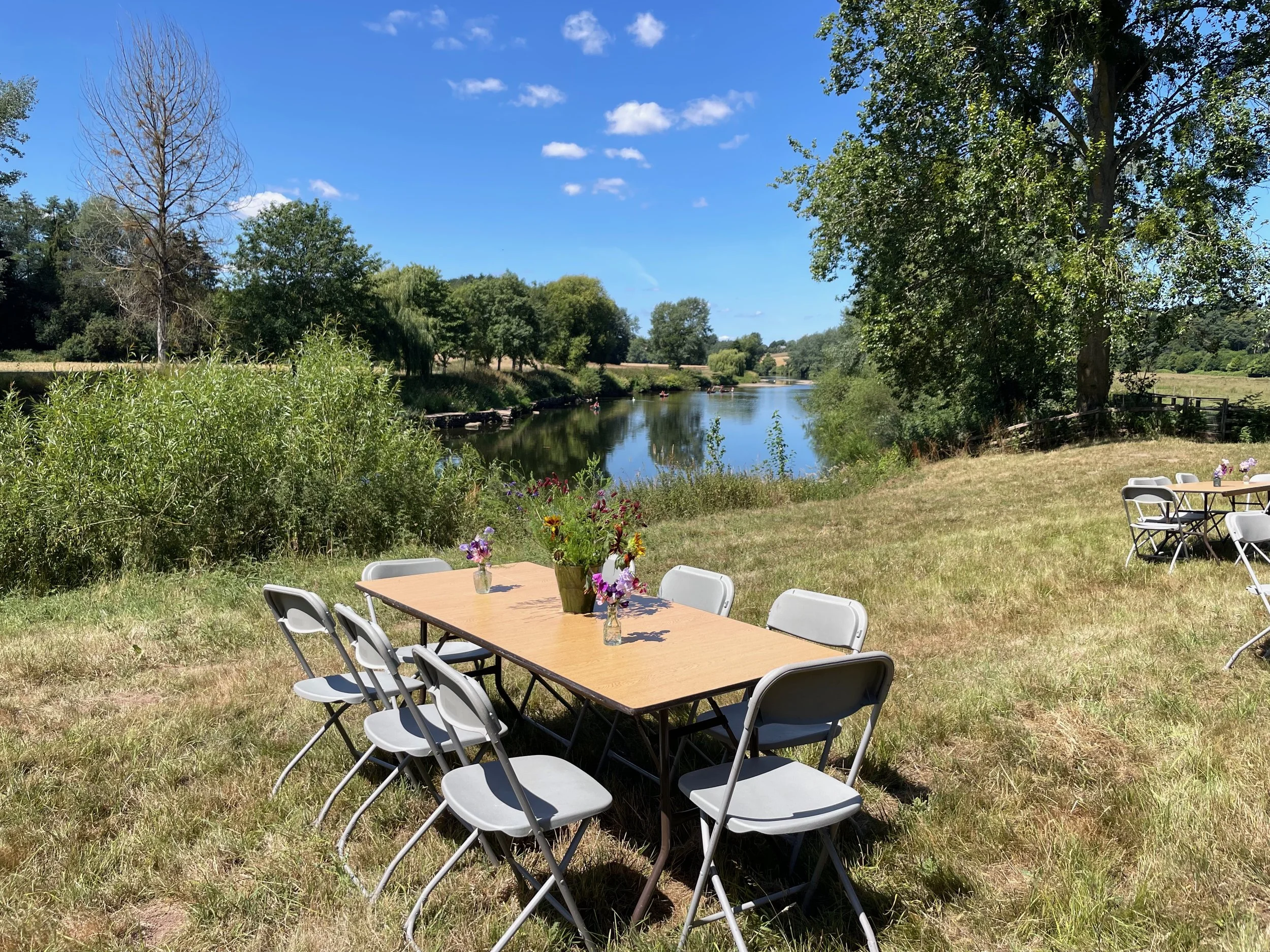 Outdoor scene with a wooden table and white folding chairs by a river, decorated with vases of colorful flowers, surrounded by green trees and grass under a blue sky with clouds.