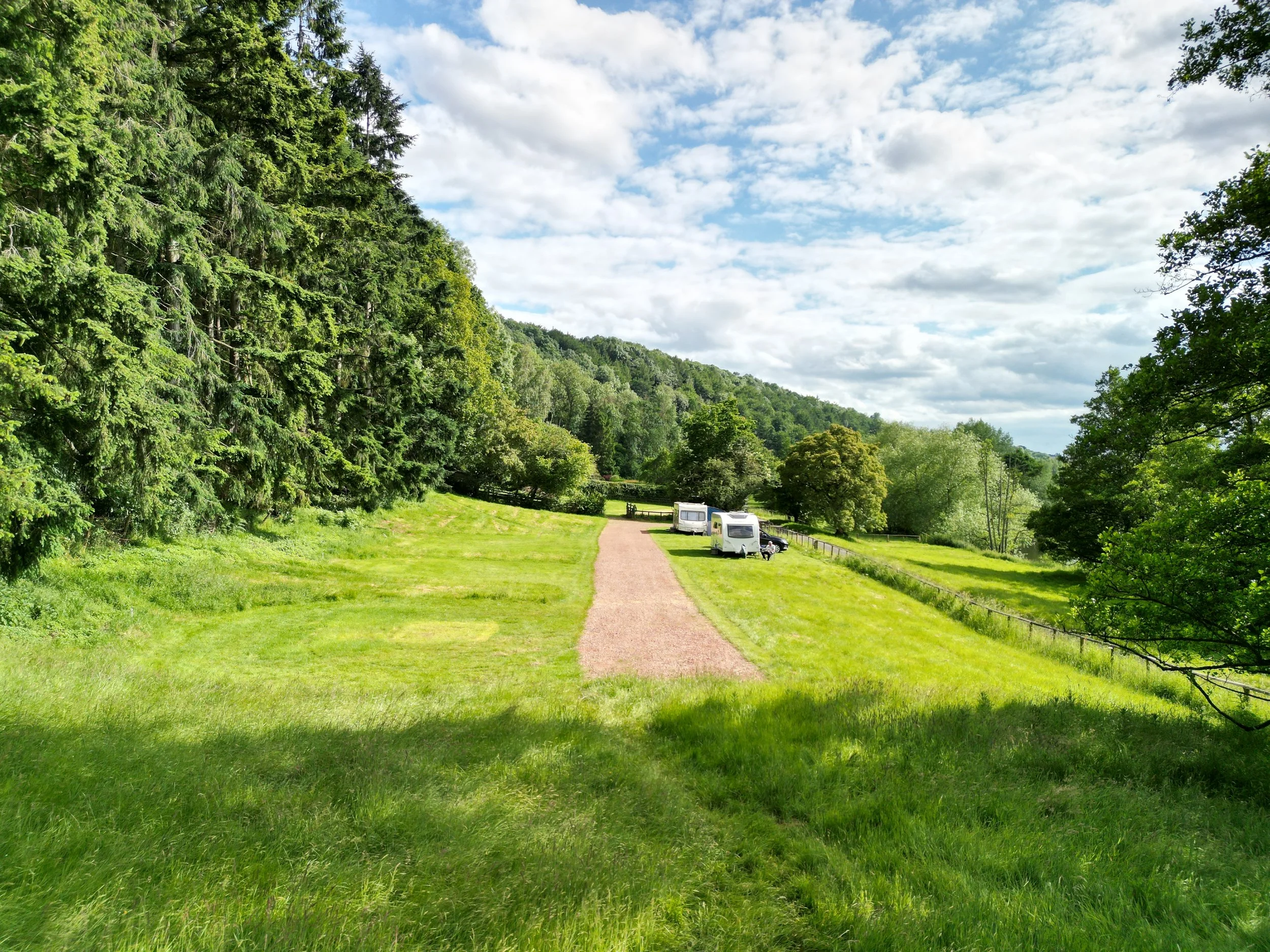 A scenic outdoor area with a dirt pathway, surrounded by lush green grass and trees, with a few small white campers parked along the right side. The sky is partly cloudy with patches of blue visible.