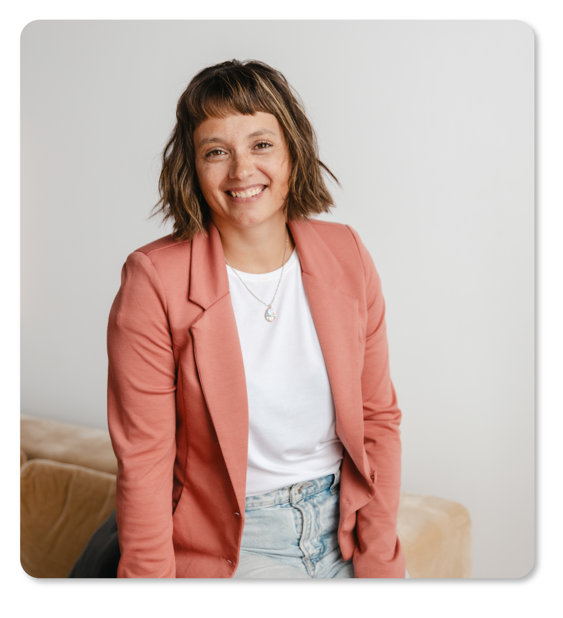 Une femme souriante avec des cheveux bruns en coiffure mi-courte, portant une veste rose, un t-shirt blanc, un collier argenté avec un pendentif, posant dans un intérieur avec un canapé beige en arrière-plan.