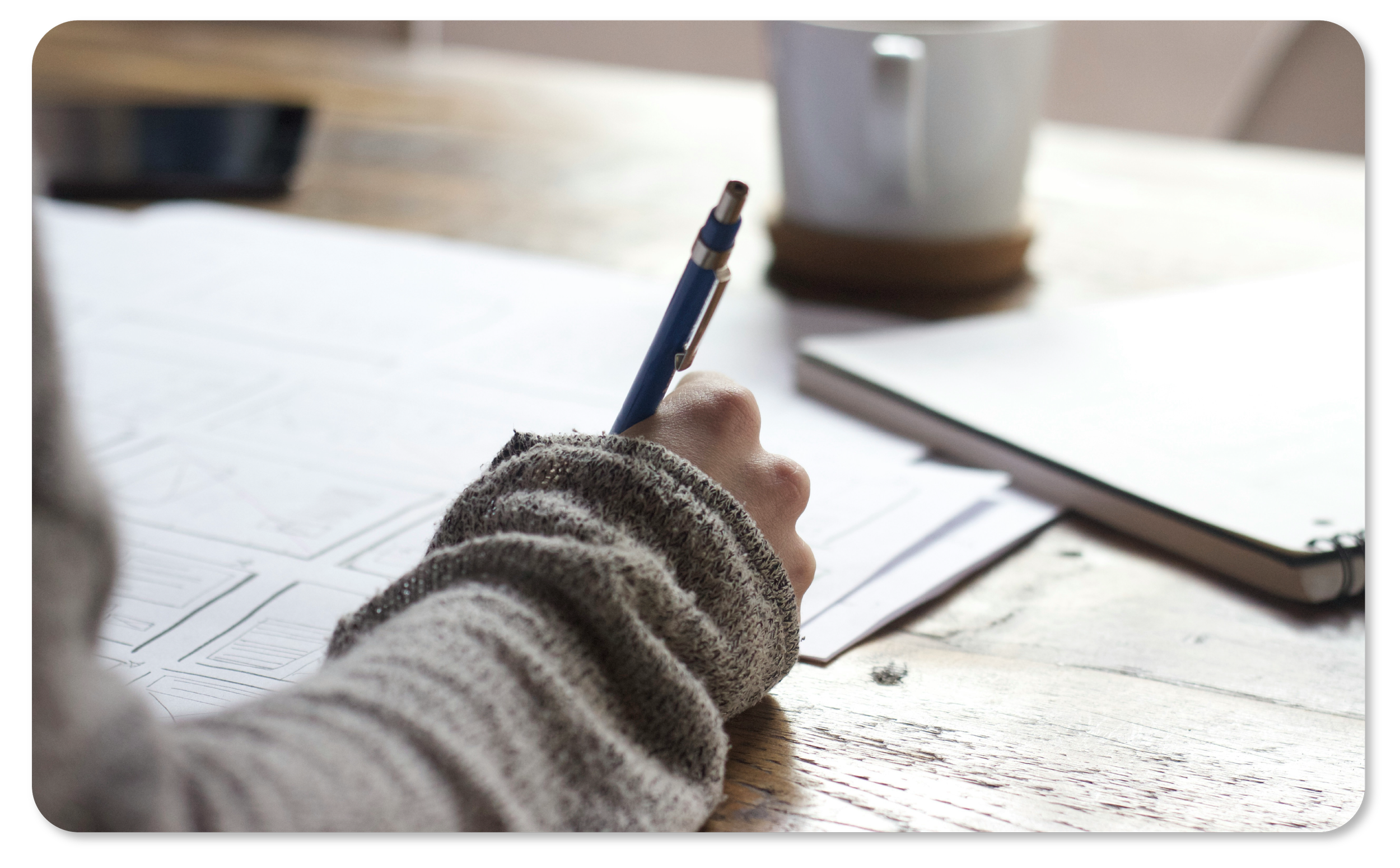 Une main d'enfant tenant un stylo bleu, écrivant ou dessinant sur des feuilles de papier avec des croquis ou des esquisses. À côté, un carnet fermé posé sur une table en bois, et une tasse blanche en arrière-plan.