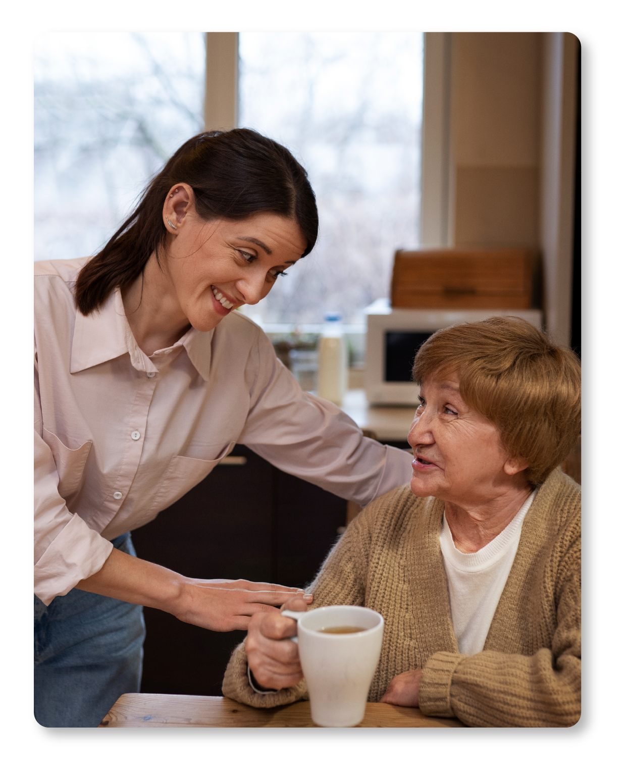 Une jeune femme sourit en parlant à une femme plus âgée qui tient une tasse de café, dans une cuisine chaleureuse.