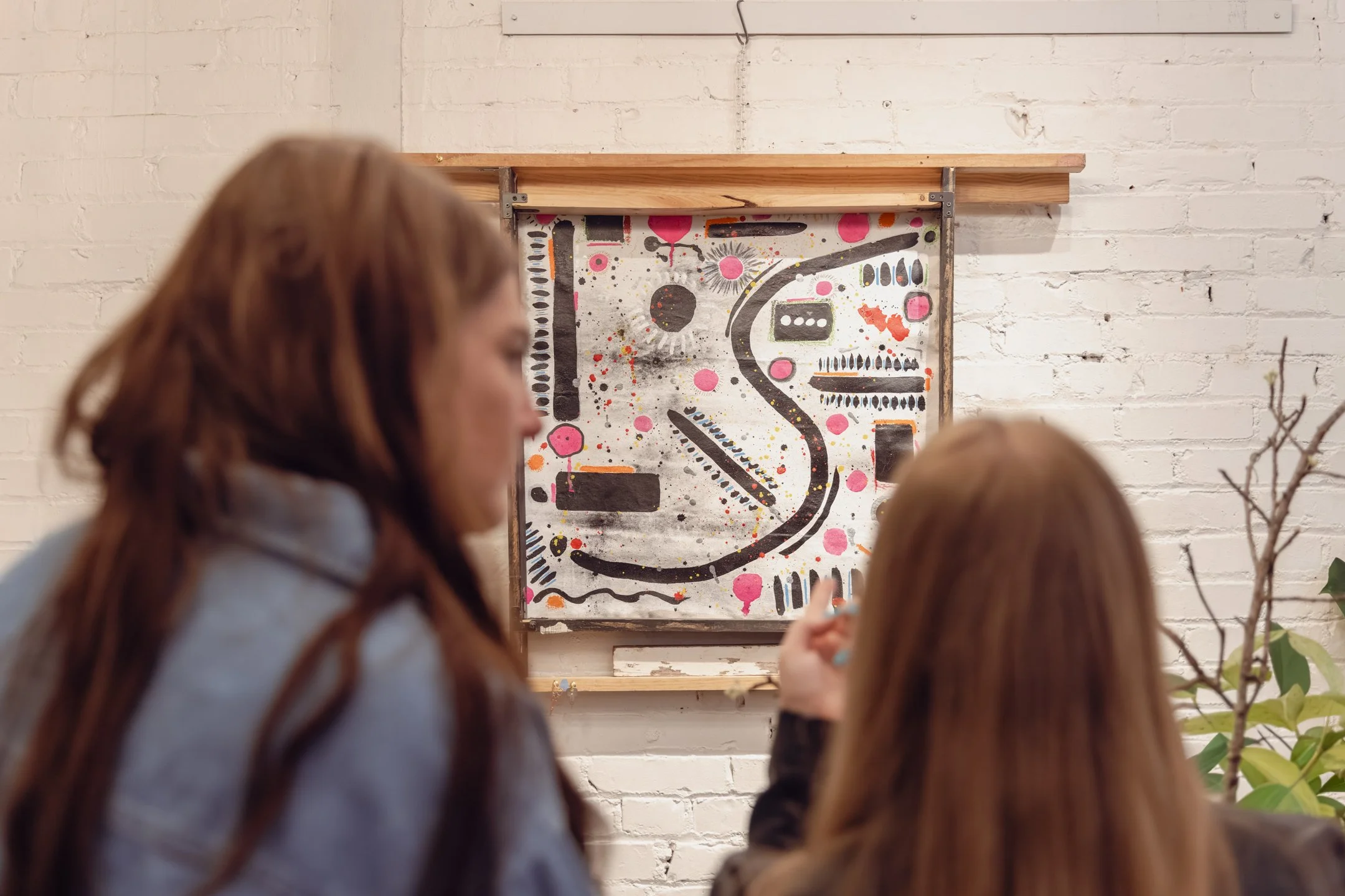 Two women with brown hair, sitting in front of abstract artwork featuring a black winding line, pink dots, and various shapes on a white brick wall.