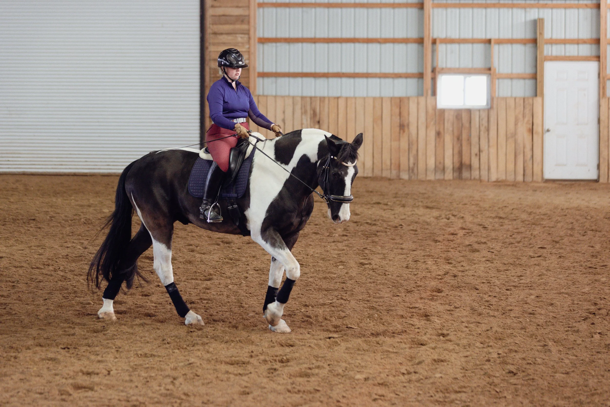 A woman riding a black and white pinto horse inside an indoor riding arena with wooden walls.