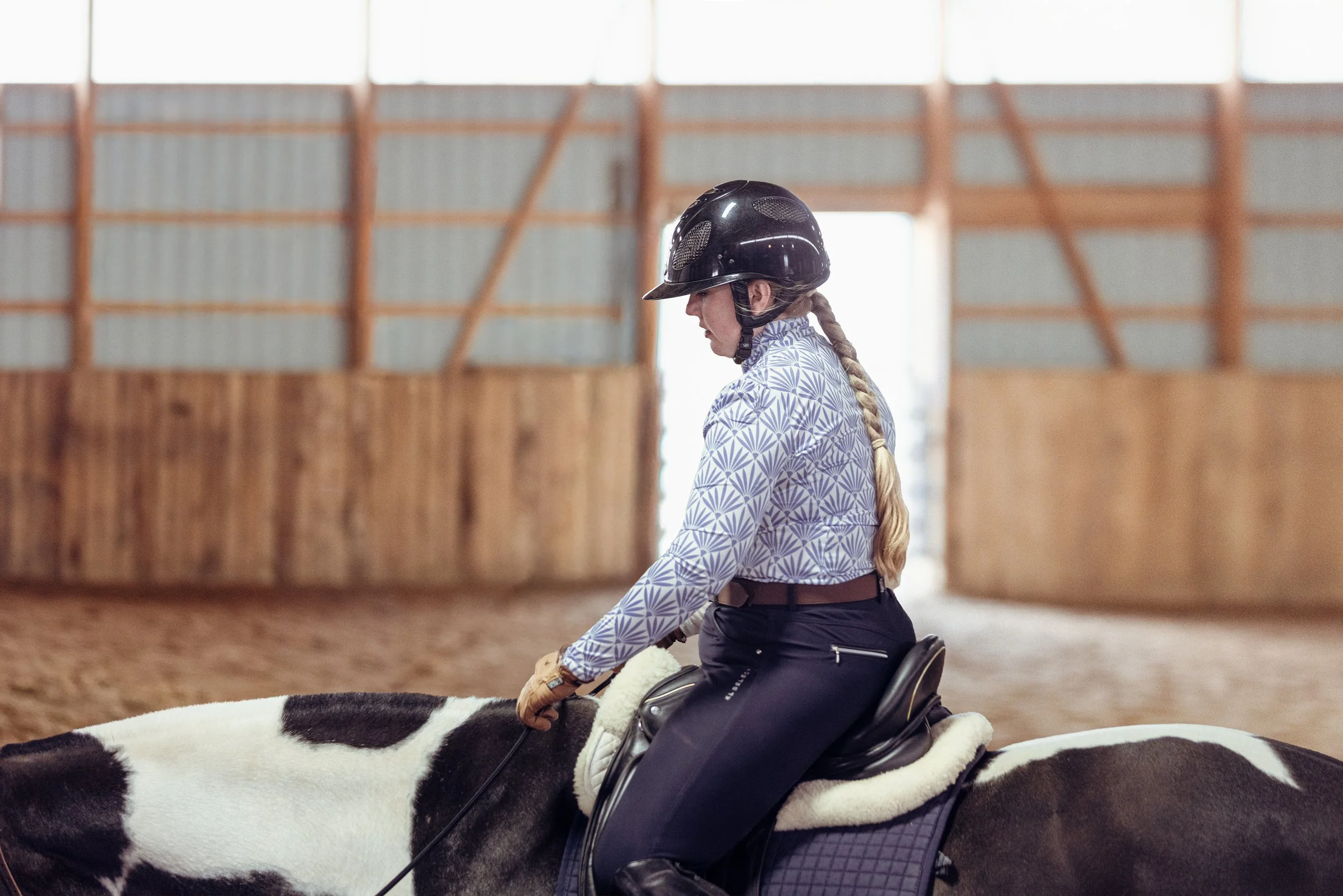 A woman riding a horse inside a wooden indoor riding arena, wearing a black helmet, patterned shirt, and riding gear.