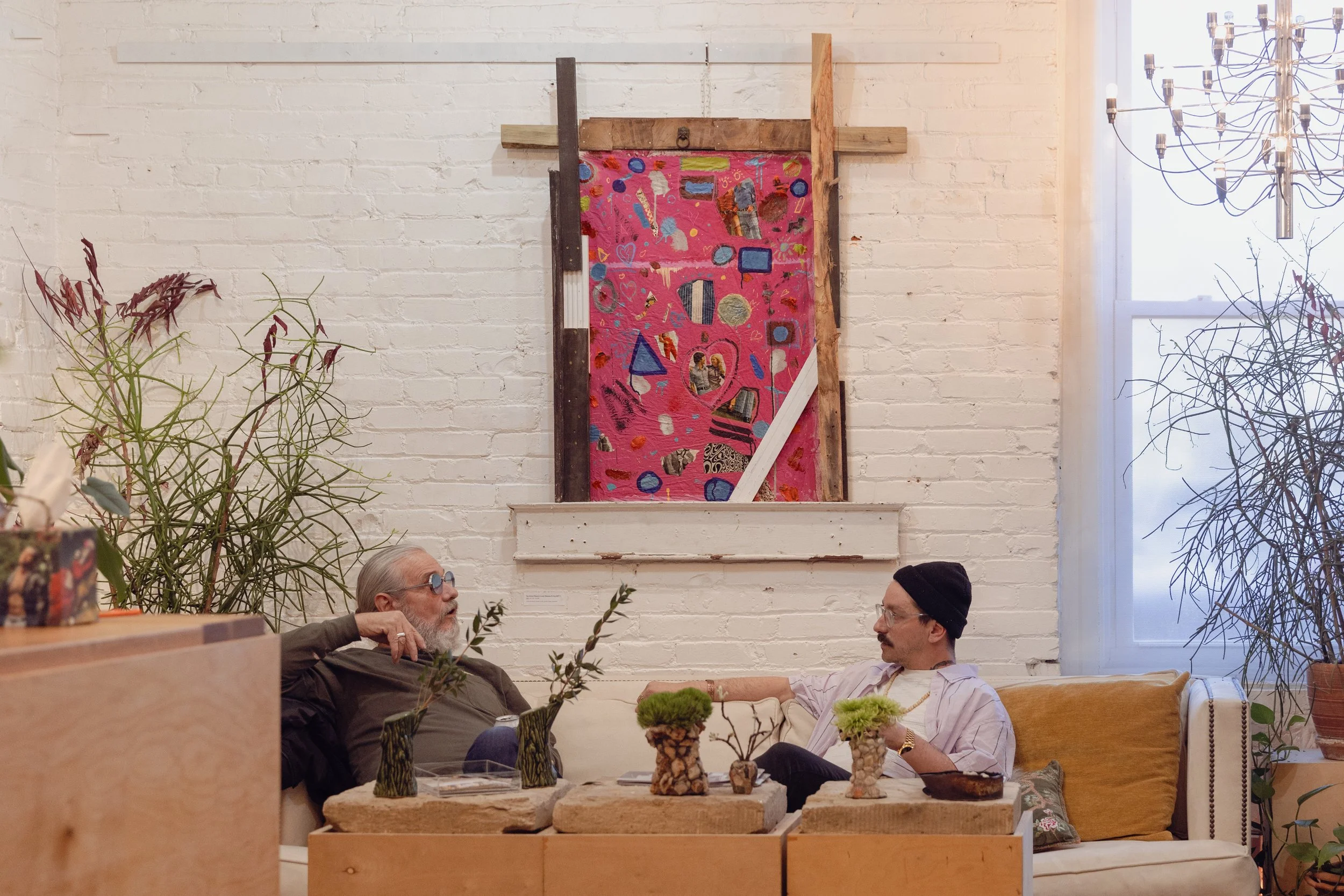 Two men are sitting and talking on a beige sofa in a art gallery or studio with white brick walls. There are various plants and vases on the table in front of them, and colorful artwork hangs on the wall behind them. One man has gray hair, a beard, and sunglasses, while the other is wearing a black beanie and glasses.