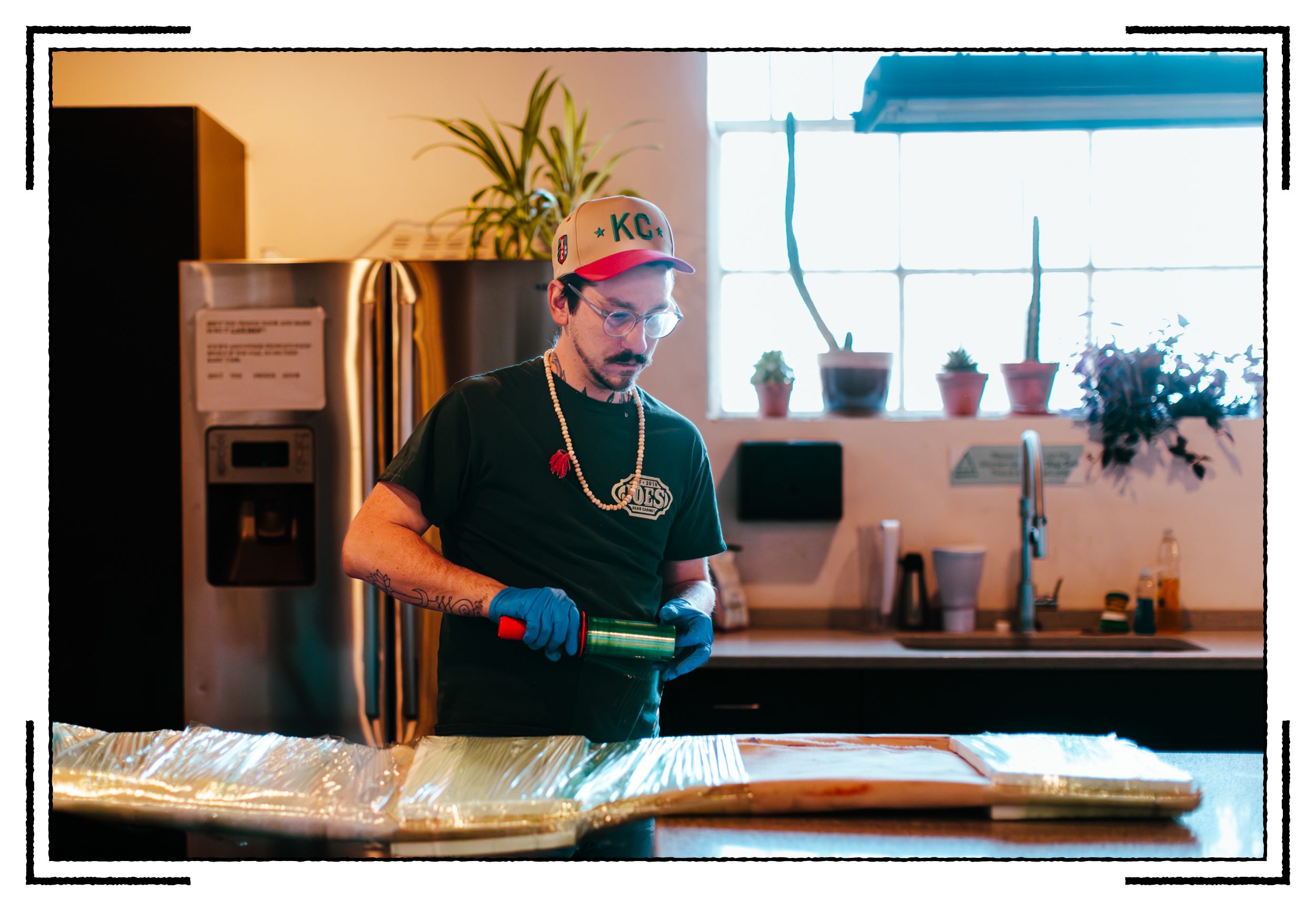 A man wearing a Kansas City Royals hat, glasses, and a black T-shirt stands in a kitchen, using a green cannoli tube to work with sheets of dough or pastry on a wooden surface covered with plastic wrap.